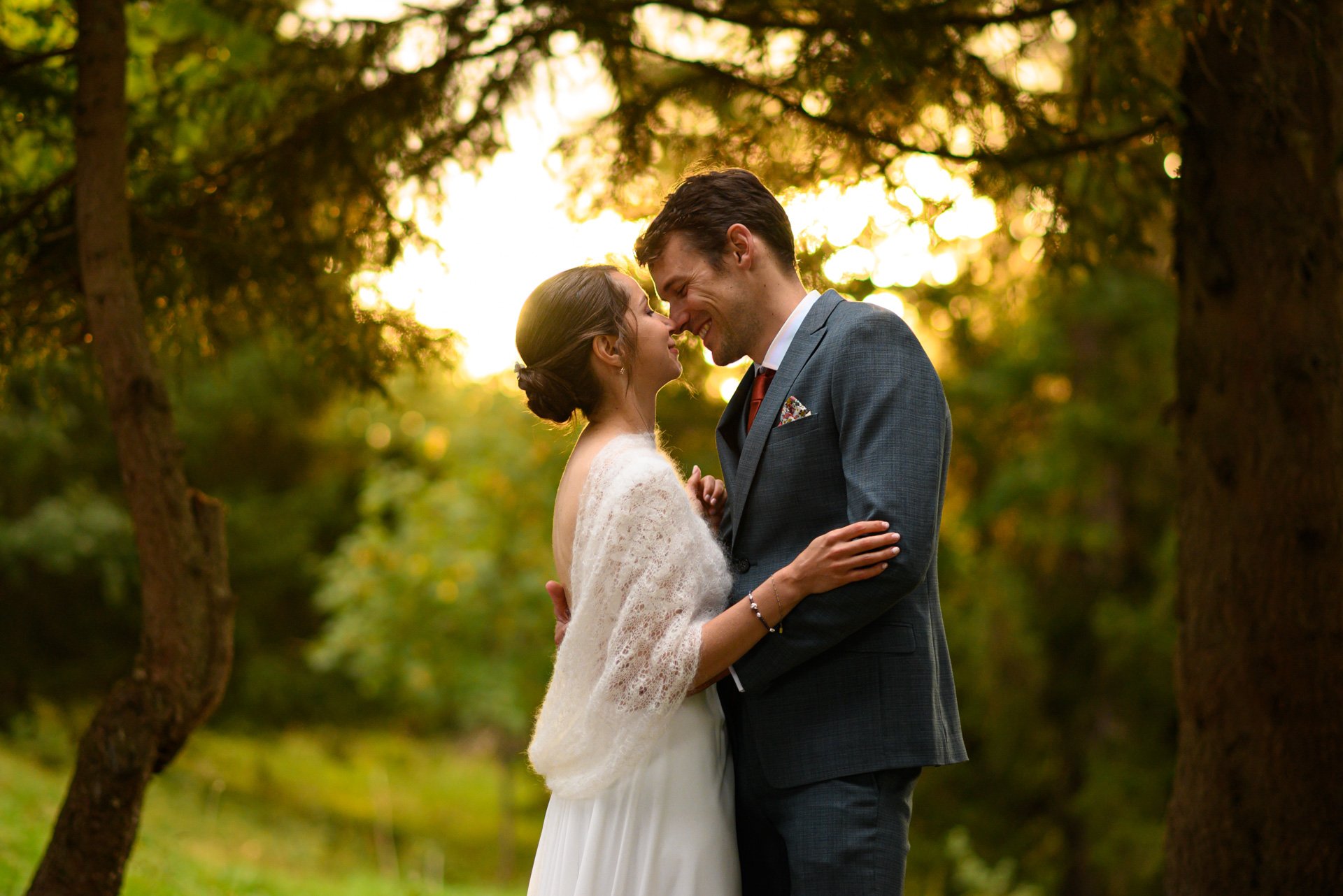 un couple de mariés souriants, debout face à face, dans une forêt lors d'un coucher de soleil