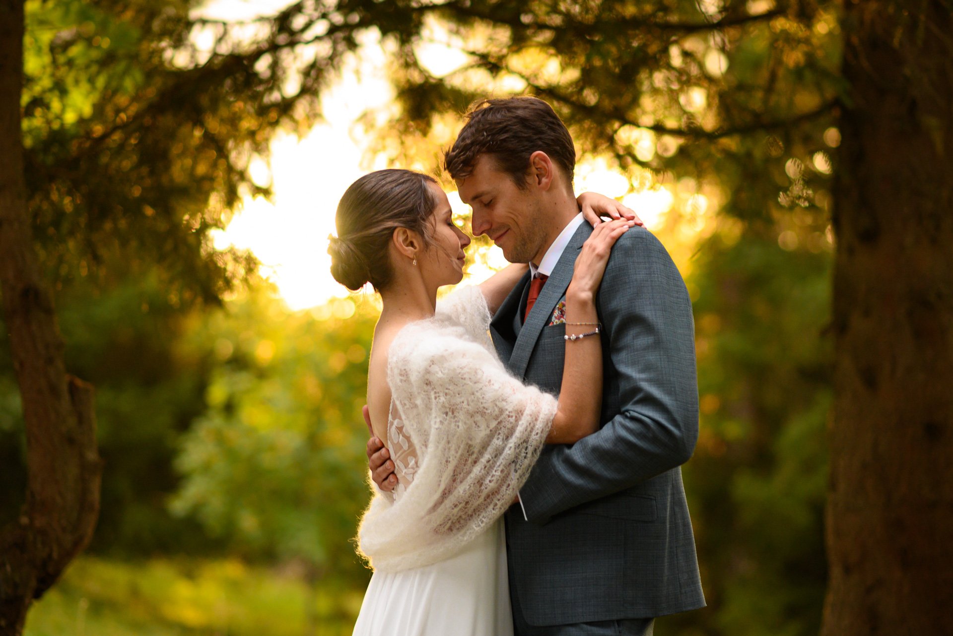 Un couple en vêtements de mariage s'enlace dans un parc, avec un arrière-plan de arbres et de lumière dorée du coucher de soleil.