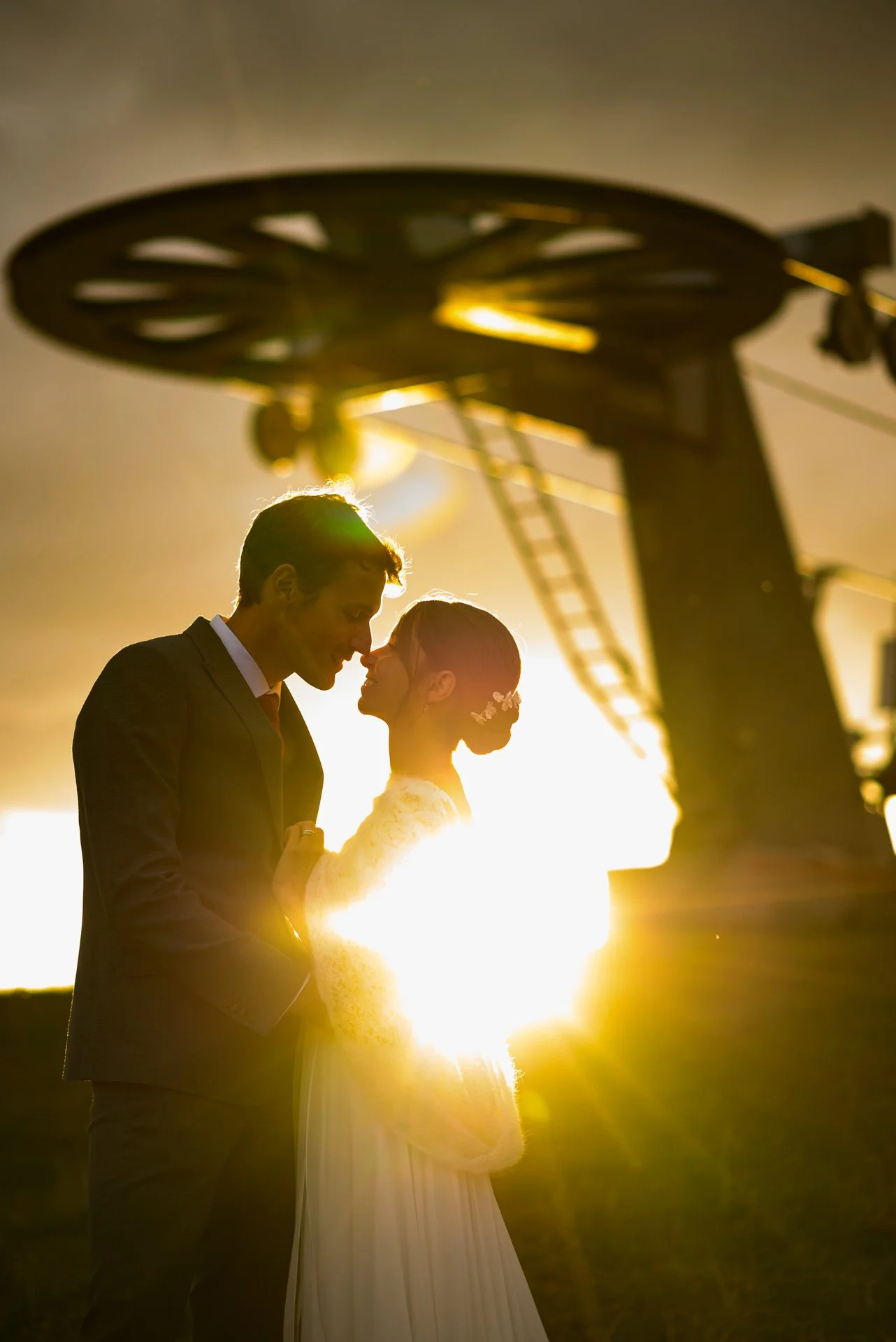 Un couple de mariés en silhouette devant un coucher de soleil, la tête proche, sous une structure de funiculaire.