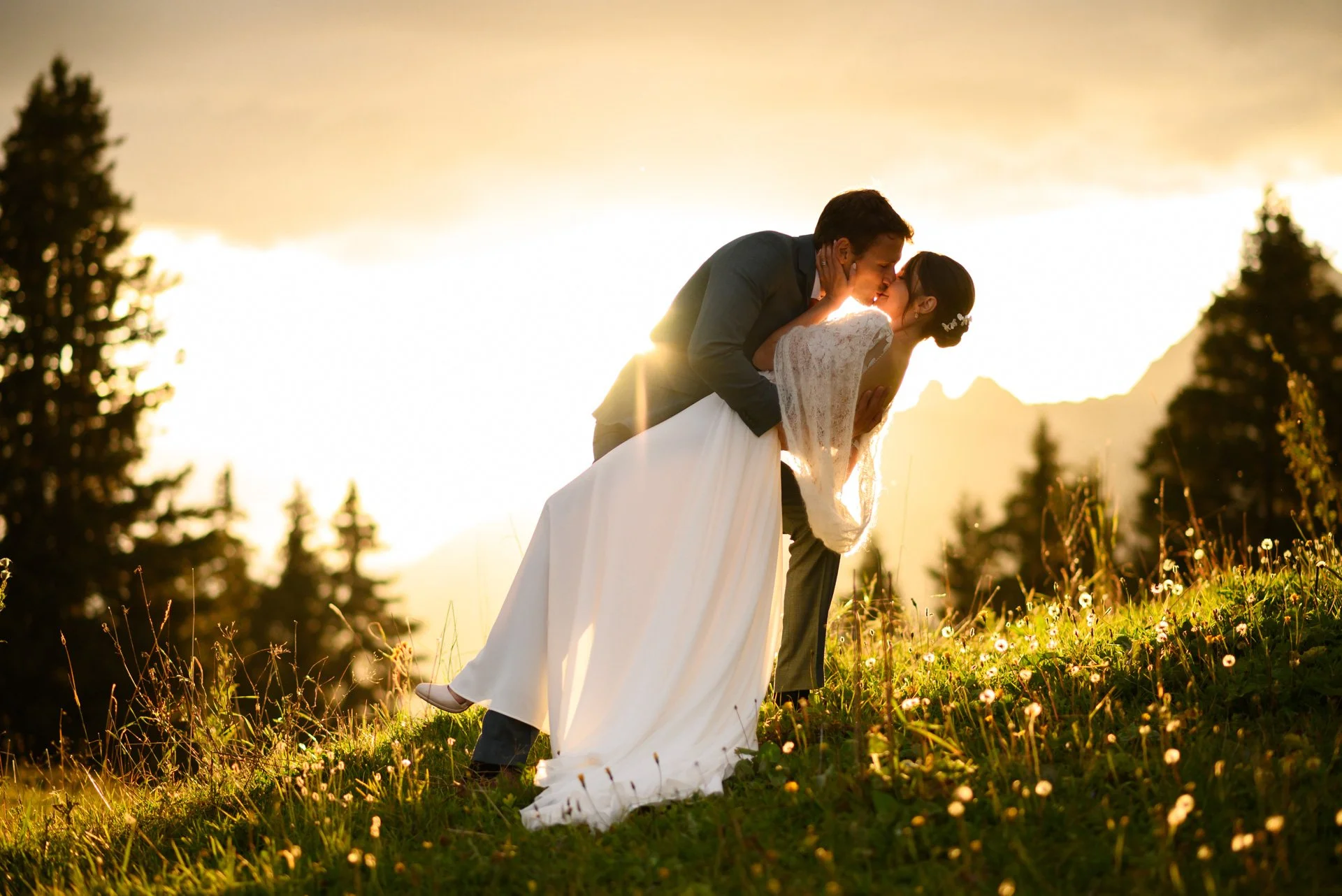 Un couple en mariage échange un baiser lors d'un coucher de soleil dans un champ, avec des montagnes en arrière-plan.