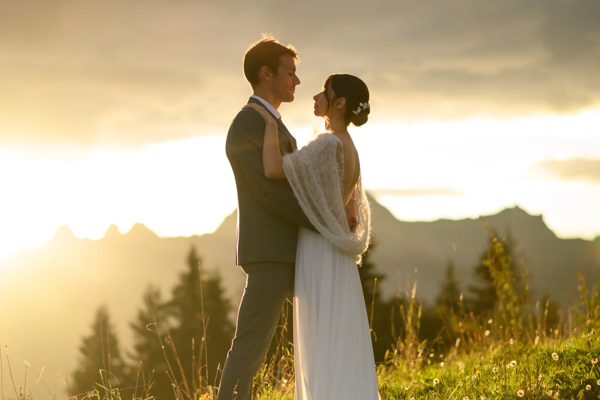 Un couple en costume de mariage s'enlace sur un champ avec un coucher de soleil en arrière-plan, montagne et arbres en silhouette.