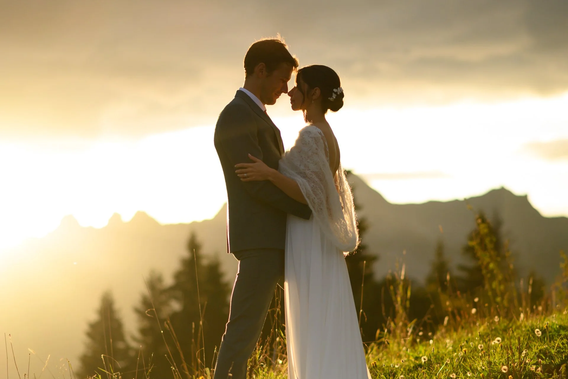 Un couple en mariage, debout dans un champ, à l'extérieur, lors d'un coucher de soleil, avec des montagnes en arrière-plan.