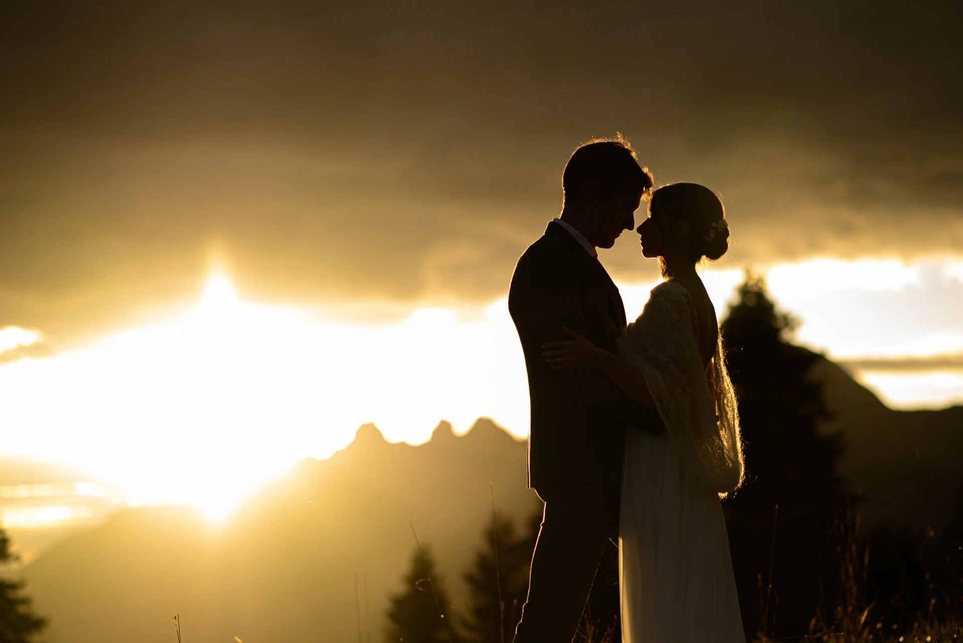 Silhouette d'un couple lors d'un mariage, se tenant face à face contre un coucher de soleil en arrière-plan, avec des montagnes et des arbres en silhouette.