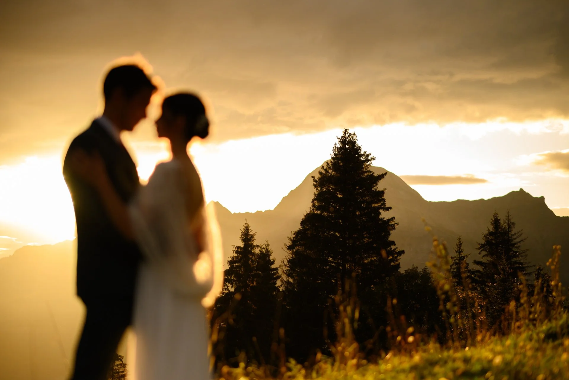 Un couple de mariés en silhouette lors d'un coucher de soleil en pleine nature, avec des montagnes et des arbres en arrière-plan.