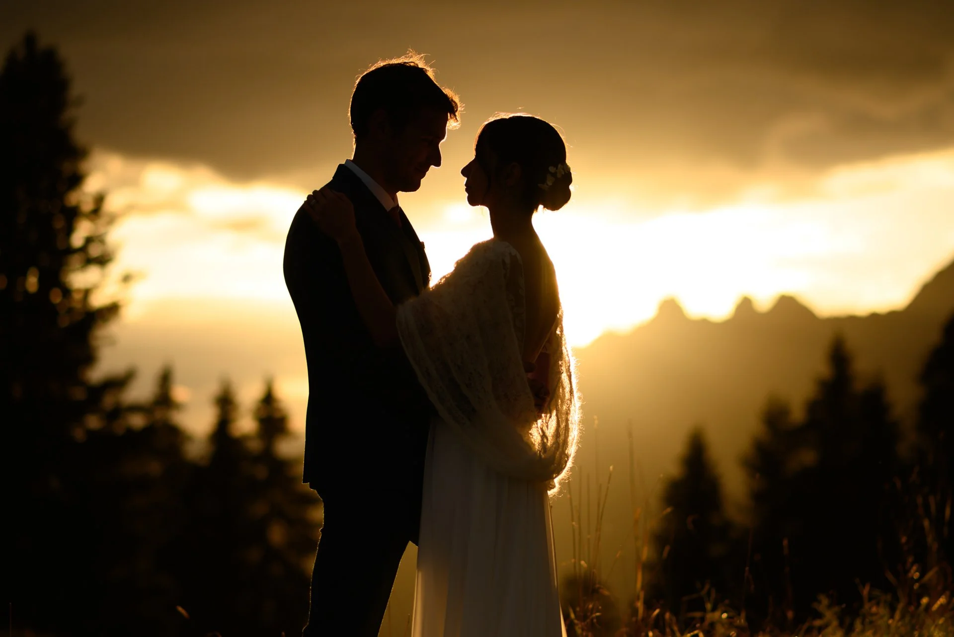Un couple de mariés regardant dans les yeux lors d'un coucher de soleil, en silhouette, avec des arbres et des montagnes en arrière-plan.