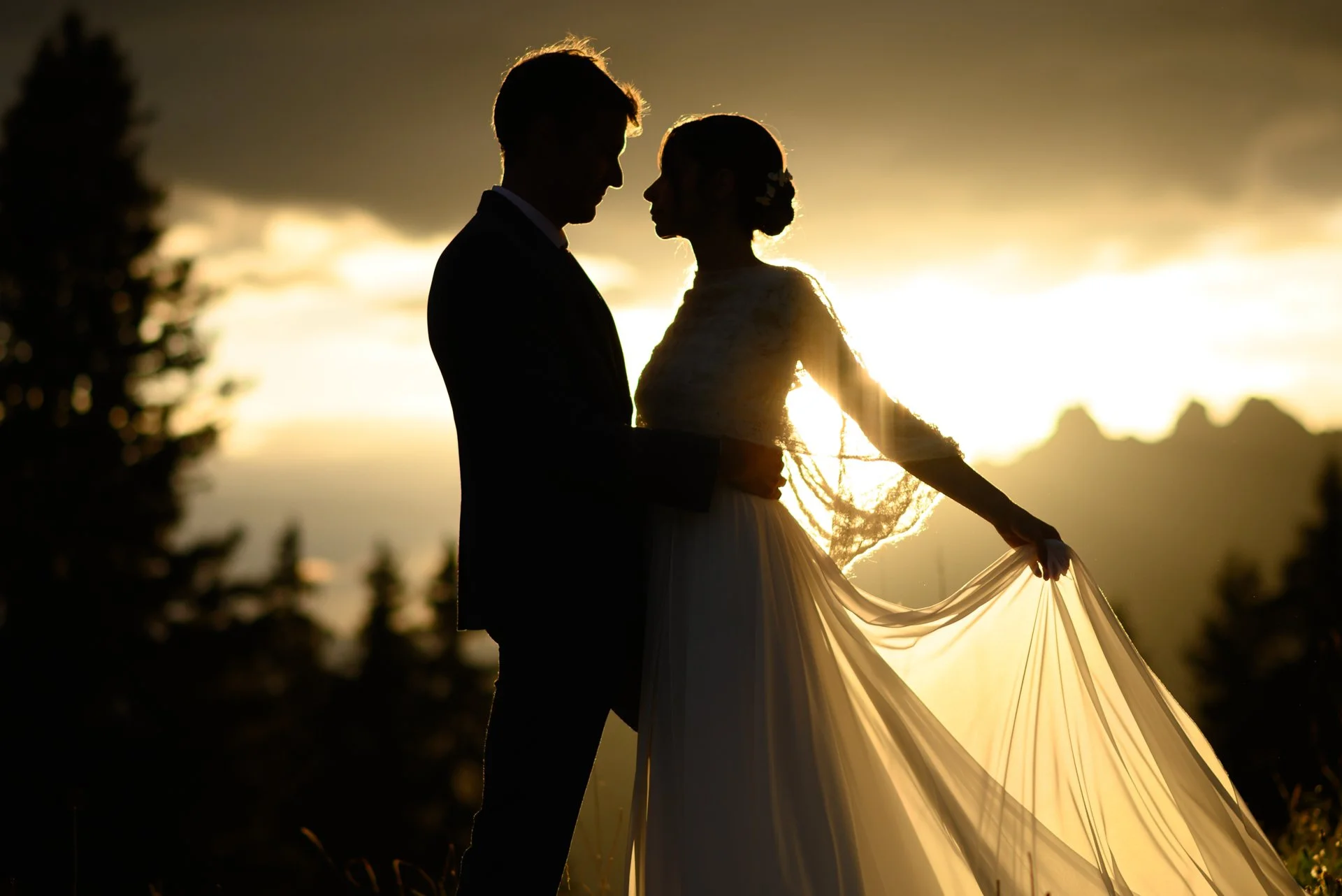 Silhouettes d'un couple de mariés lors d'un coucher de soleil, la femme portant une robe de mariée longue et le homme en costume, dans un cadre naturel avec des montagnes en arrière-plan.