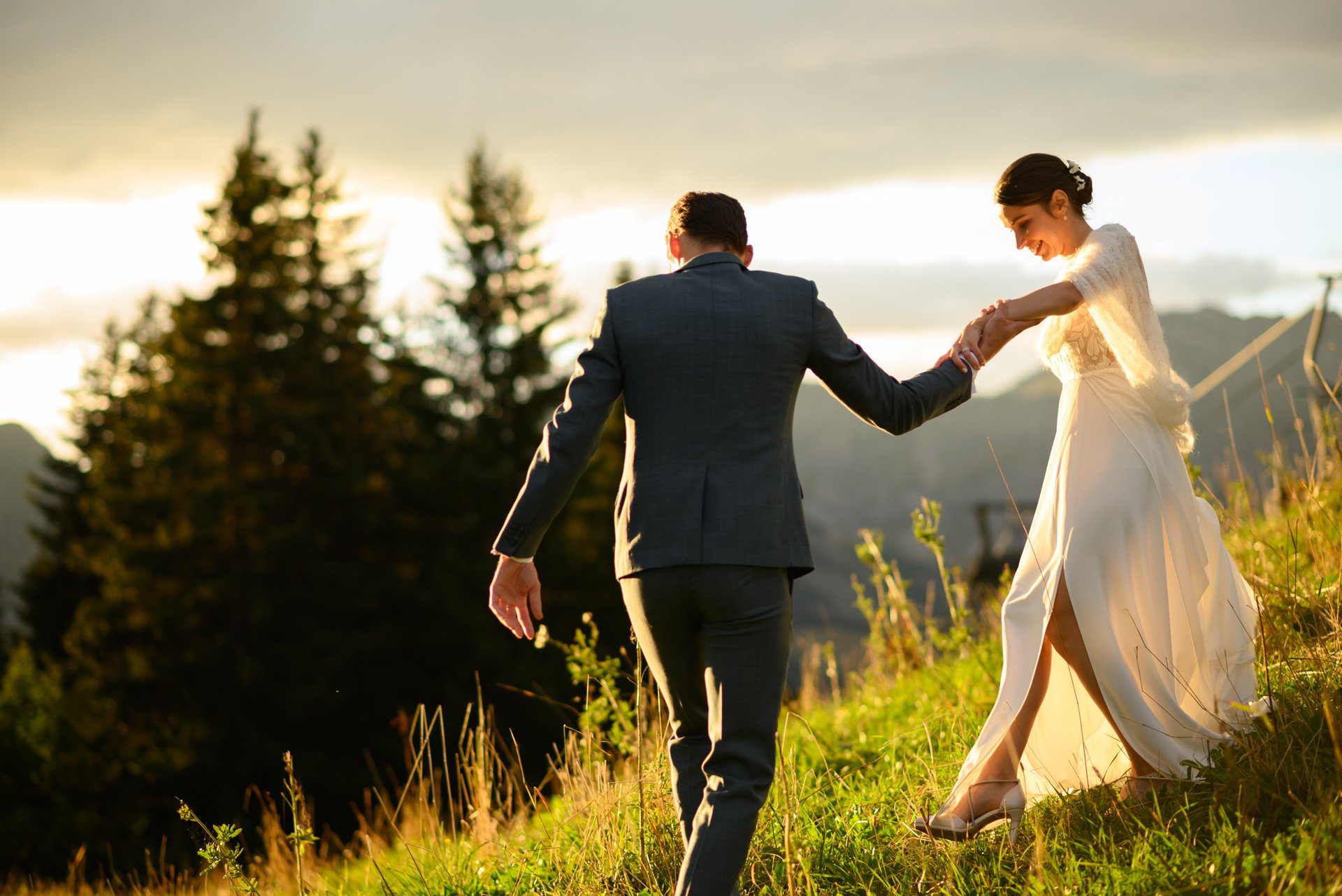Un couple en tenue de mariage marche dans une prairie lors d'un coucher de soleil, avec des arbres en arrière-plan.
