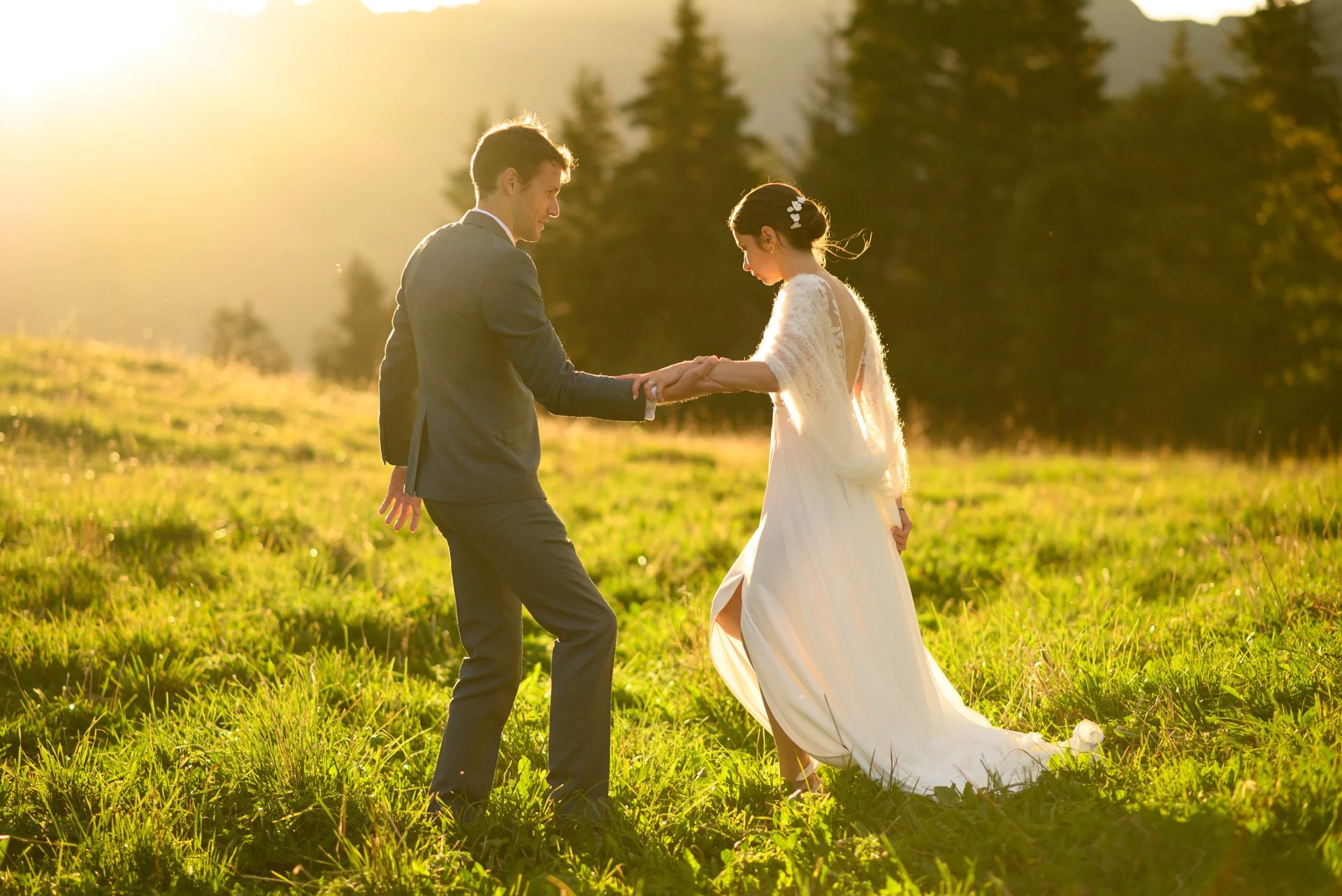 Un couple en tenue de mariage danse dans un champ en plein air au coucher du soleil, entouré d'arbres.