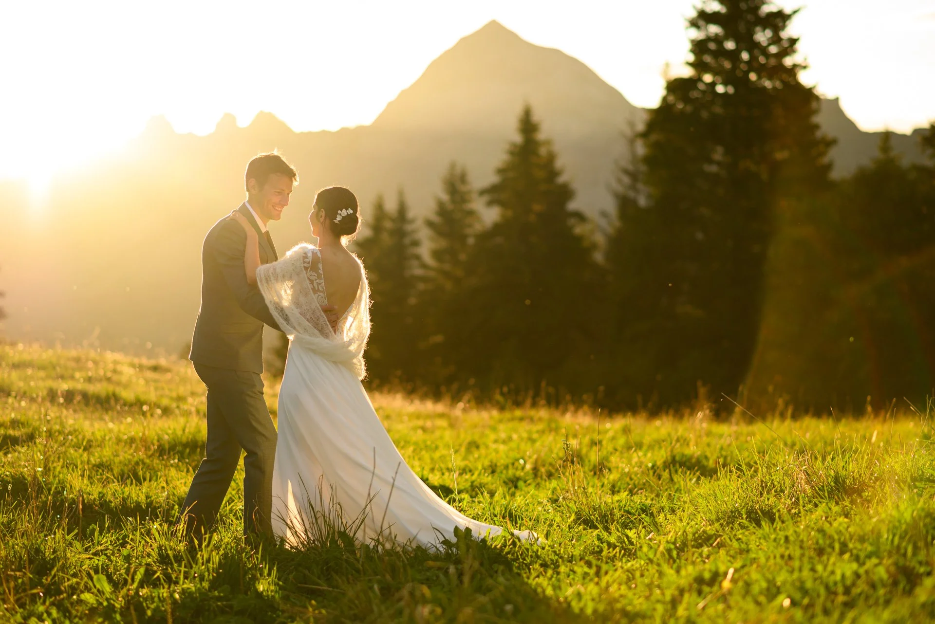 Un couple de mariés danse dans un champ lors d'un coucher de soleil, avec des montagnes et des arbres en arrière-plan.