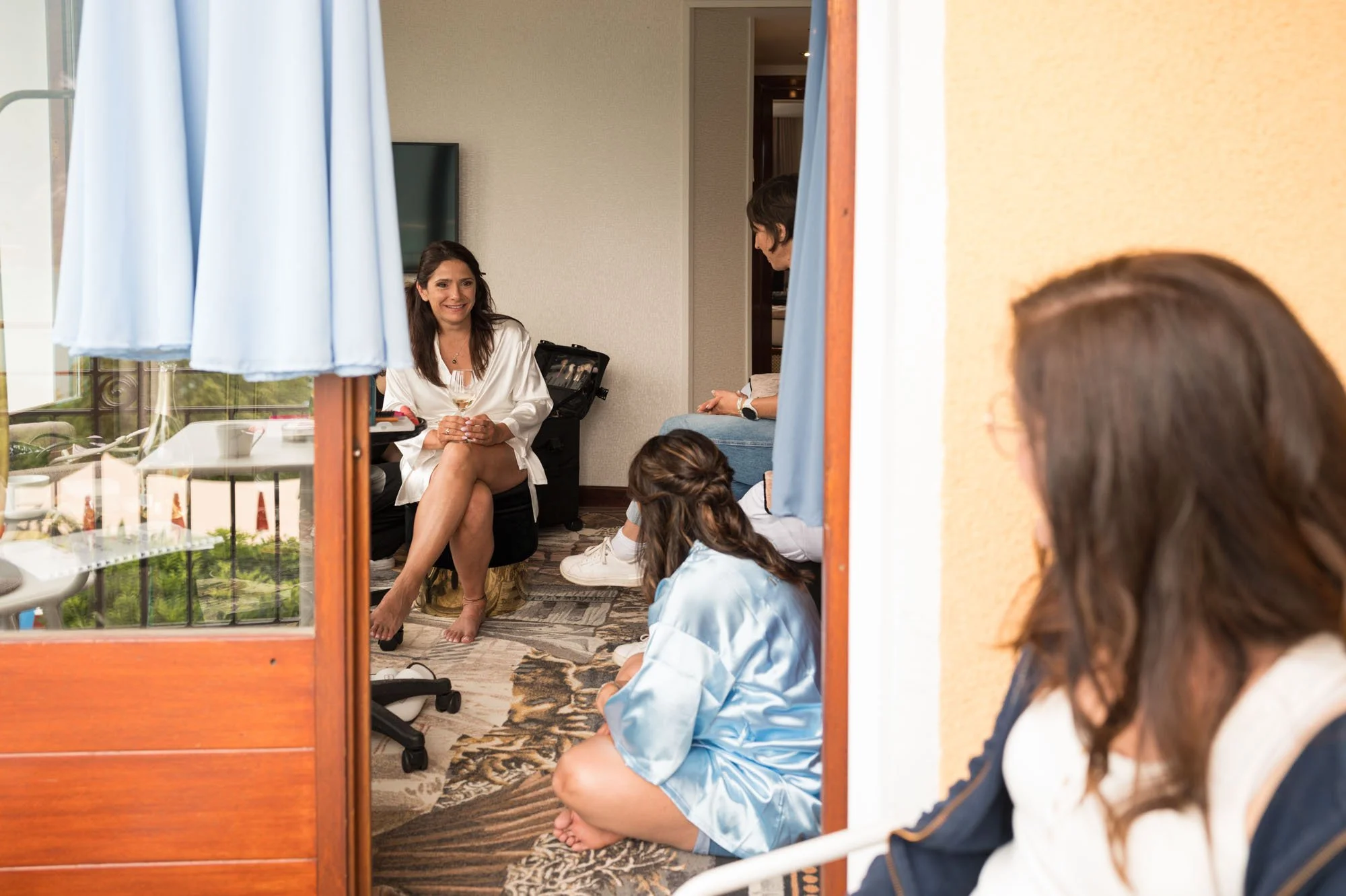 Femme assise avec un verre de vin, entourée de jeunes femmes dans une chambre d'hôtel.