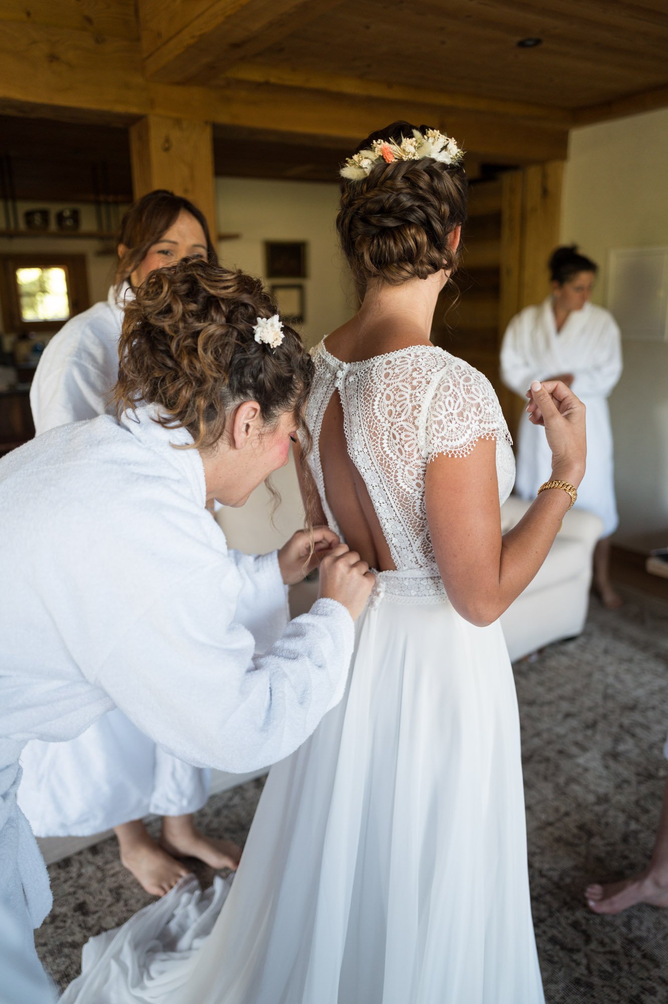 Jeune femme en robe de mariée avec dos en dentelle, entourée de ses demoiselles d'honneur préparant la robe