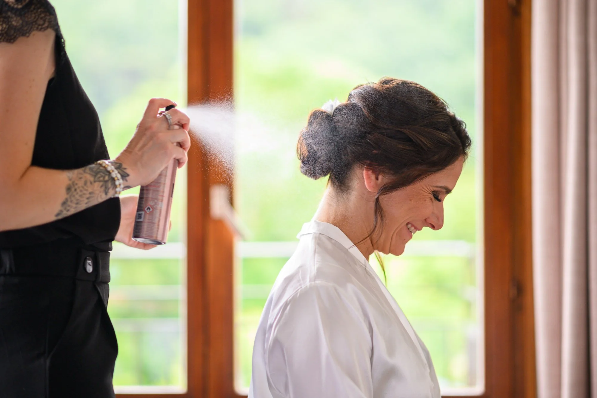 Une femme maquille une autre femme avec un spray pendant que celle-ci sourit, assise devant une grande fenêtre avec un cadre en bois.