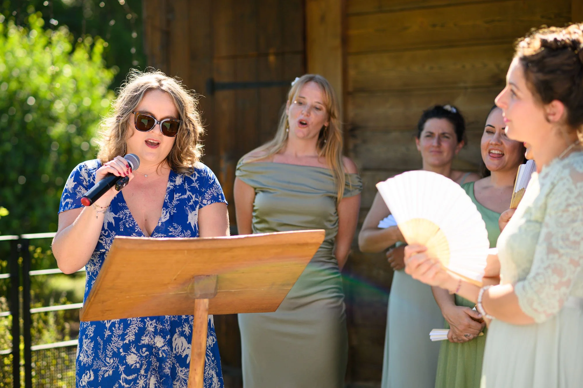Groupe de femmes lors d'une cérémonie en extérieur, une femme en bleu chantant ou récitant en lisant un texte, avec d'autres femmes en arrière-plan.