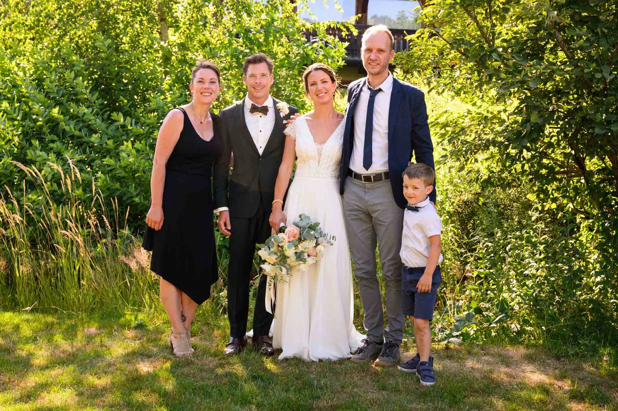 Groupe de six personnes lors d'un mariage en plein air, avec une femme en robe de mariée blanche tenant un bouquet, entourée de deux hommes en costume, une femme en robe noire et un jeune garçon en chemise blanche et bermuda bleu.
