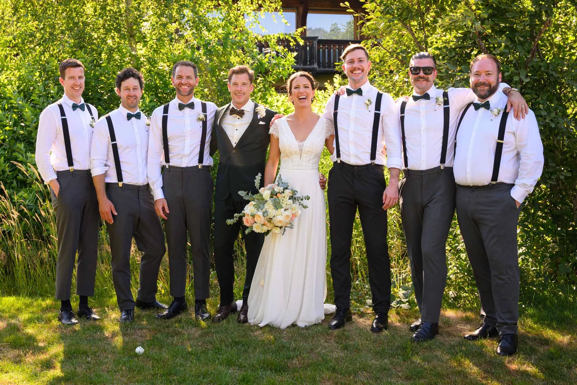 Groupe de huit personnes lors d'un mariage, posant dehors en été avec des arbres en arrière-plan, dont la mariée en robe blanche avec bouquet de fleurs, et le marié en costume noir. Les autres hommes portent une chemise blanche avec un pantalon gris 