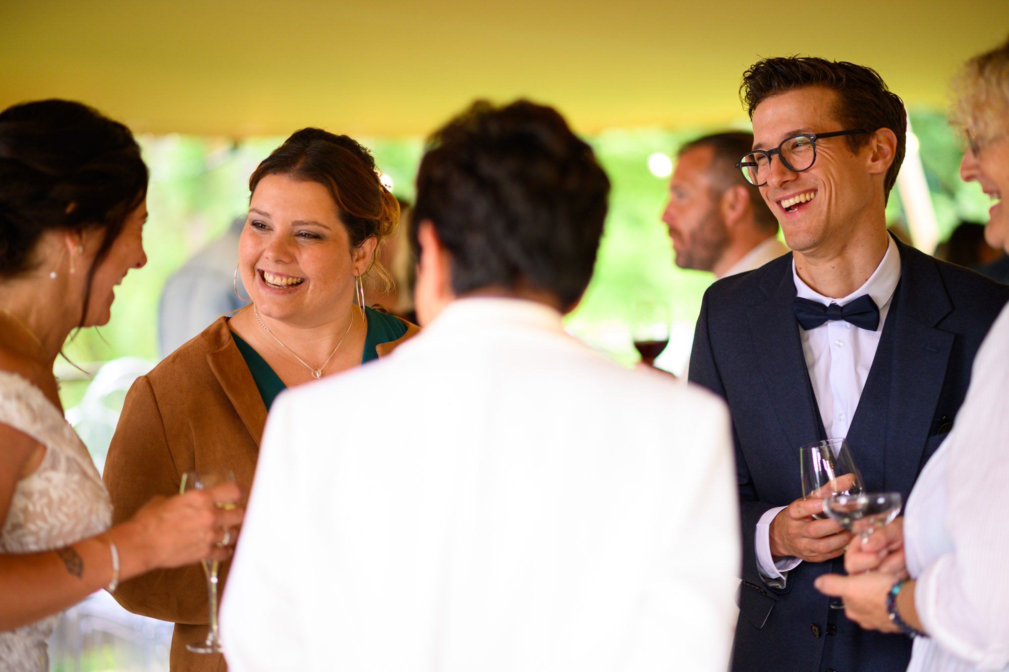 Groupe de personnes lors d'une célébration, souriant, tenant des verres, en tenue élégante, en extérieur avec une tente et un fond verdoyant.