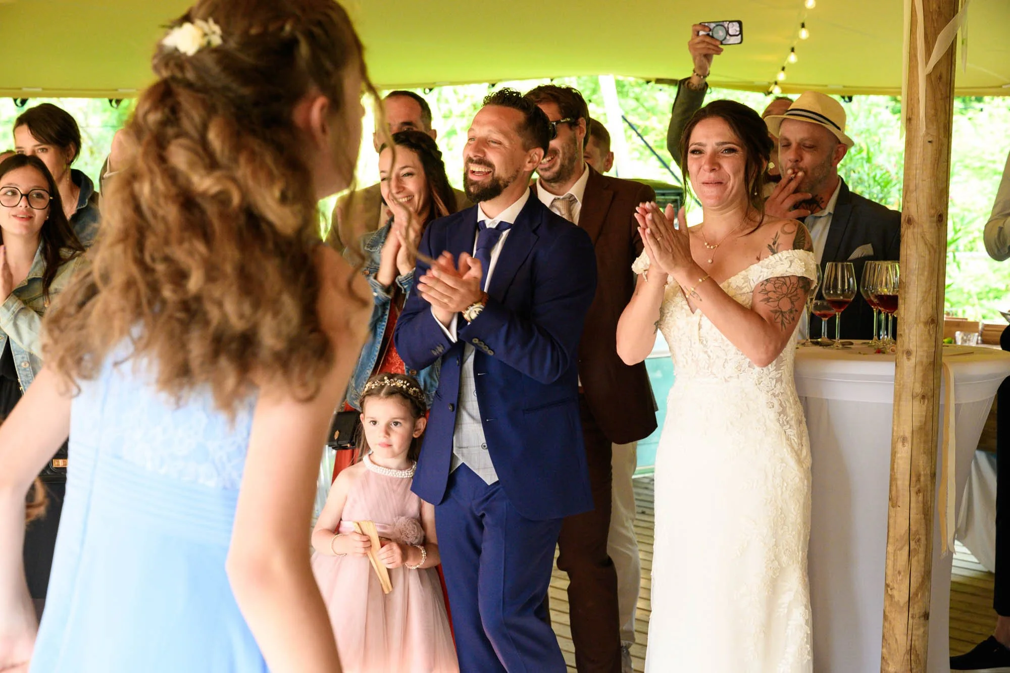 Un groupe de personnes lors d'un mariage, applaudissant et souriant, avec la mariée en robe blanche dans le cadre chaleureux d'une tente en bois.