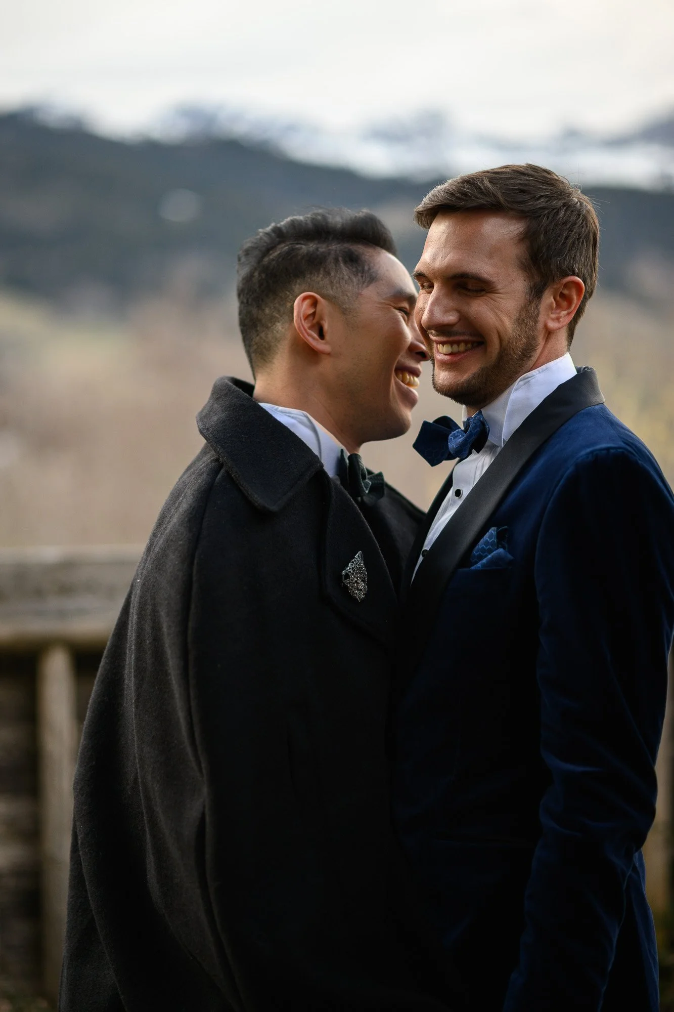 Deux hommes habillés en tuxedo, souriants, en tain de danser ou de se faire un câlin en extérieur avec un paysage de montagnes en arrière-plan.