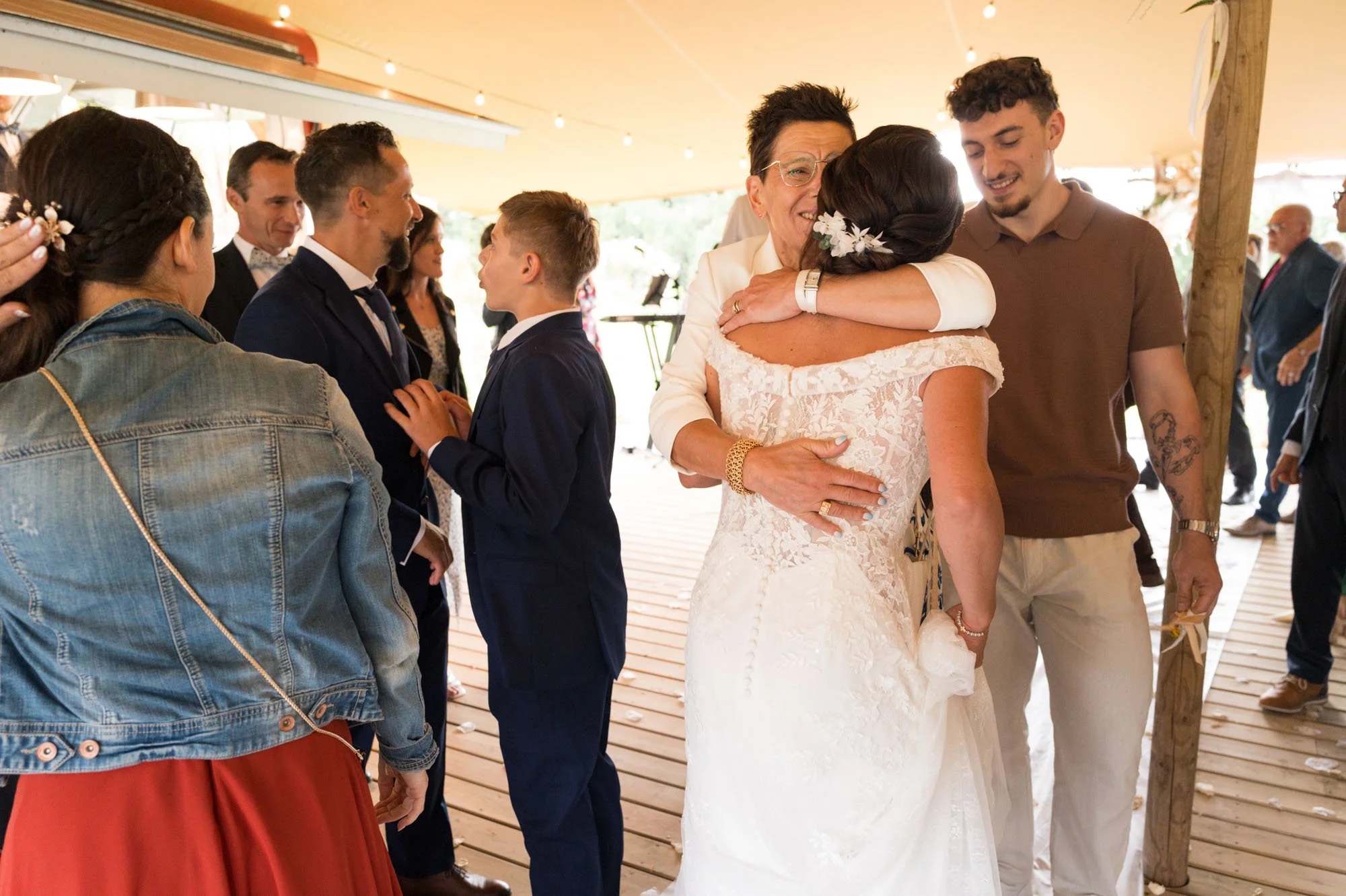 Deux femmes en robes de mariée s'embrassent, entourées de plusieurs personnes, lors d'une célébration de mariage en plein air sous un chapiteau.