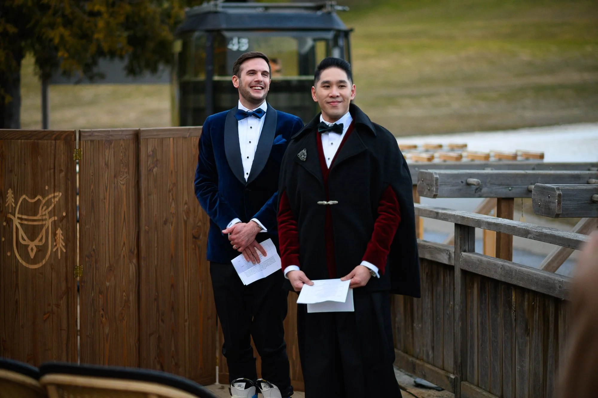 Deux hommes habillés en tenue formelle, un en smoking bleu et l'autre en manteau noir avec des accents rouges, se tiennent dans un cadre extérieur avec un plan d'eau et une construction en bois en arrière-plan.
