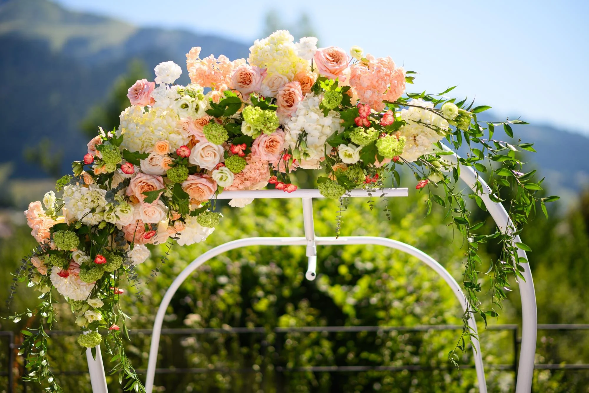 Une décoration florale de mariage avec des fleurs roses, blanches, et vertes sur une arche blanche, en extérieur avec un fond de montagnes et de végétation.