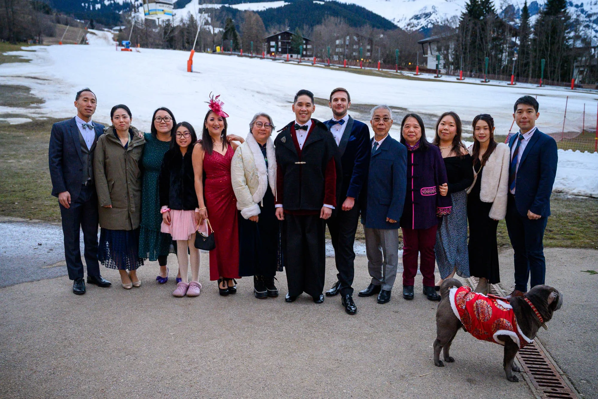 Groupe de personnes habillées élégamment posant à l'extérieur avec une station de ski en arrière-plan, et un chien portant un manteau rouge.
