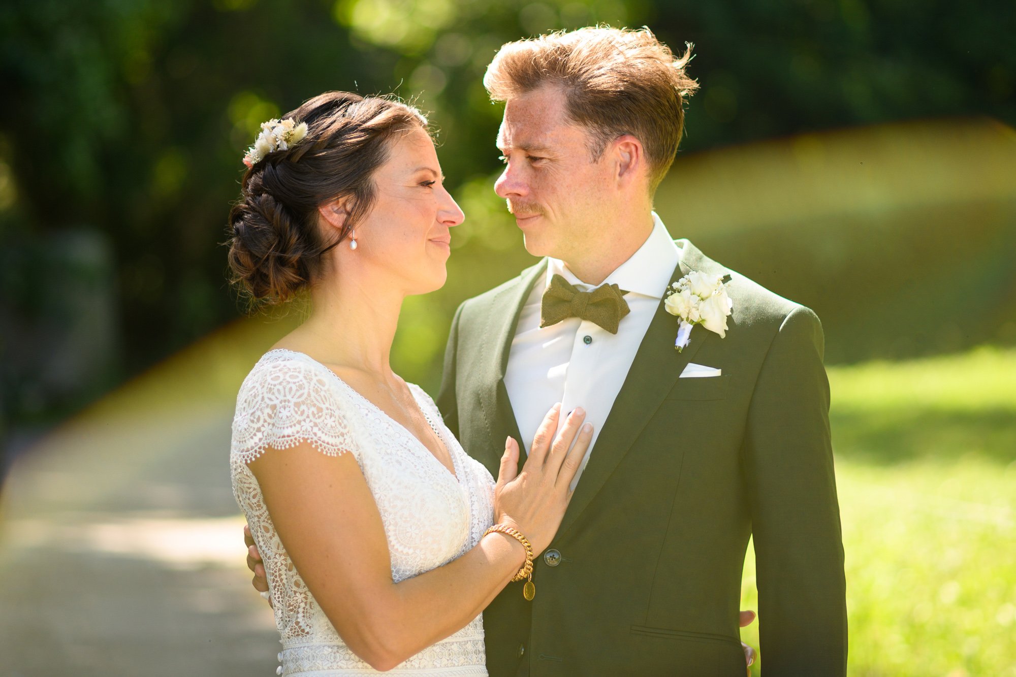 Un couple de mariage en plein air, la femme en robe blanche avec une coiffure tressée et fleurs, l'homme en smoking avec nœud papillon, se regardant tendrement.