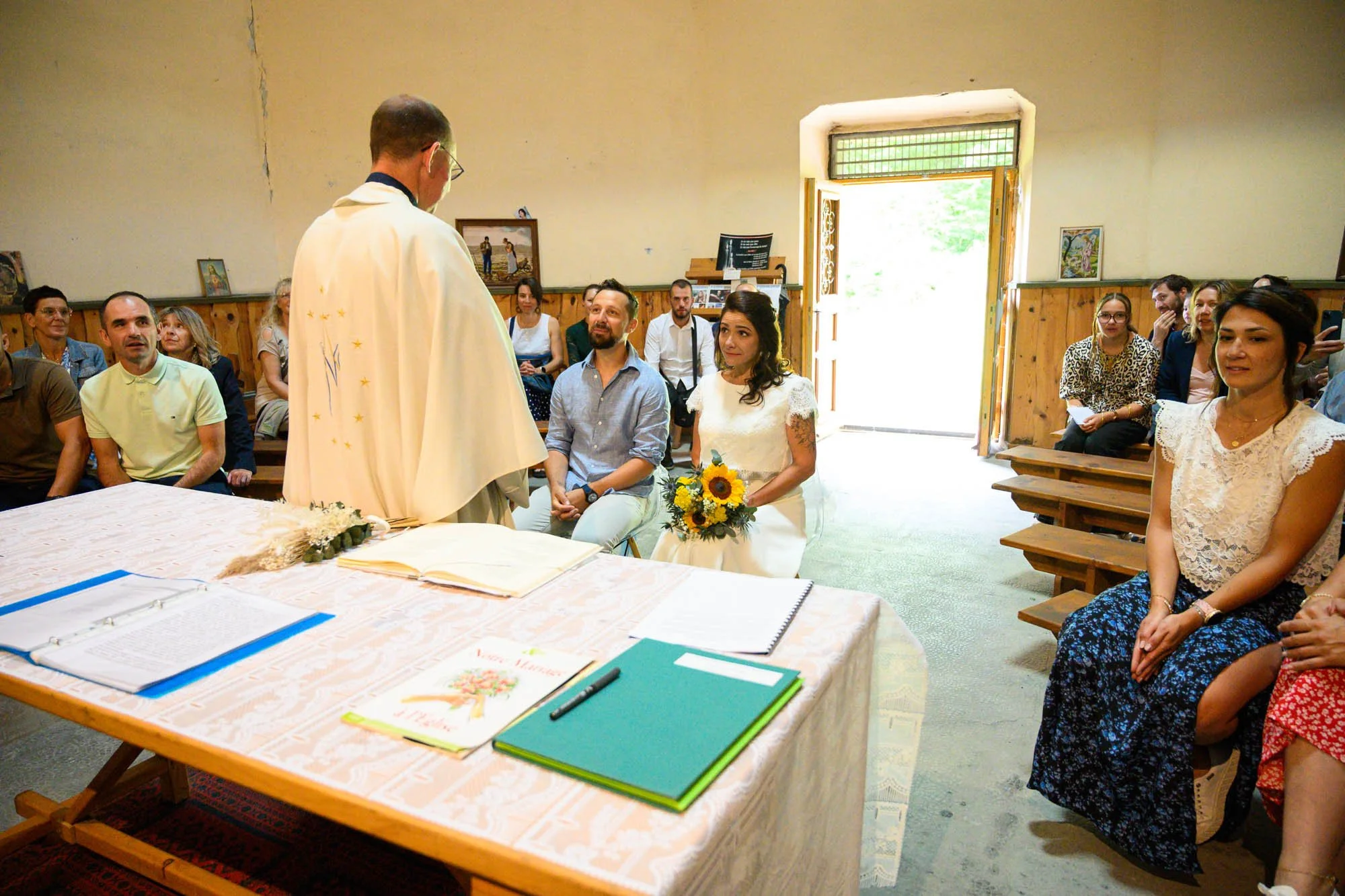 Un mariage civil en intérieur avec le prêtre ou officiant en robe blanche demandant à la mariée et au marié des vœux. La mariée porte une robe blanche et tient un bouquet de tournesols. Plusieurs invités regardent l'événement, assis sur des bancs en 
