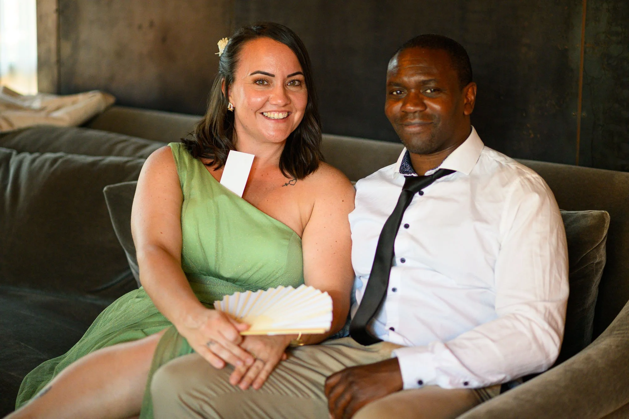 Une femme en robe verte et un homme en chemise blanche avec cravate noire assis sur un canapé, souriant.