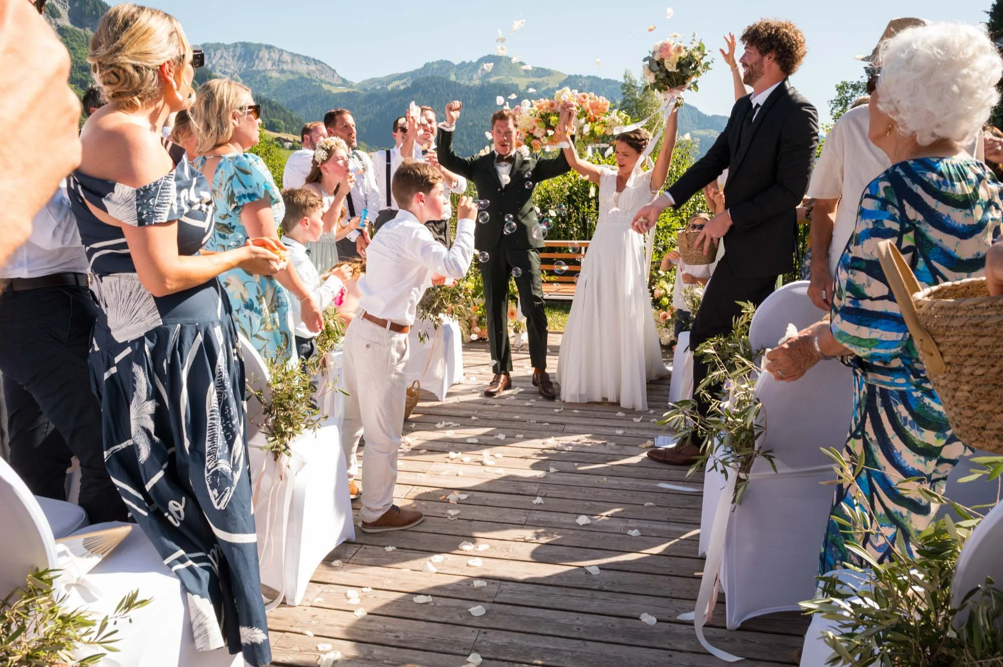 Un mariage en plein air avec un couple de mariés au centre, entouré de famille et amis qui lancent des pétales et applaudissent, en montagne par une journée ensoleillée.