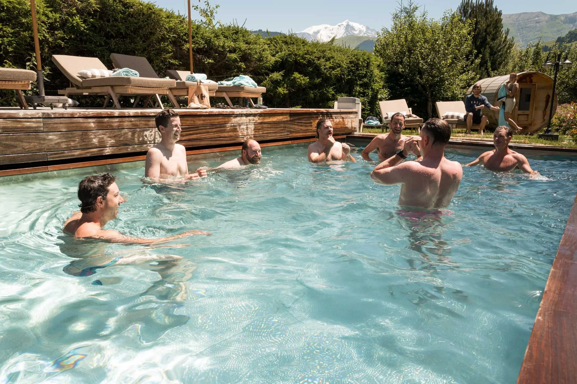 Groupe d'hommes nageant et discutant dans une piscine extérieure ensoleillée, entourée de chaises longues et de nature avec des montagnes enneigées en arrière-plan.