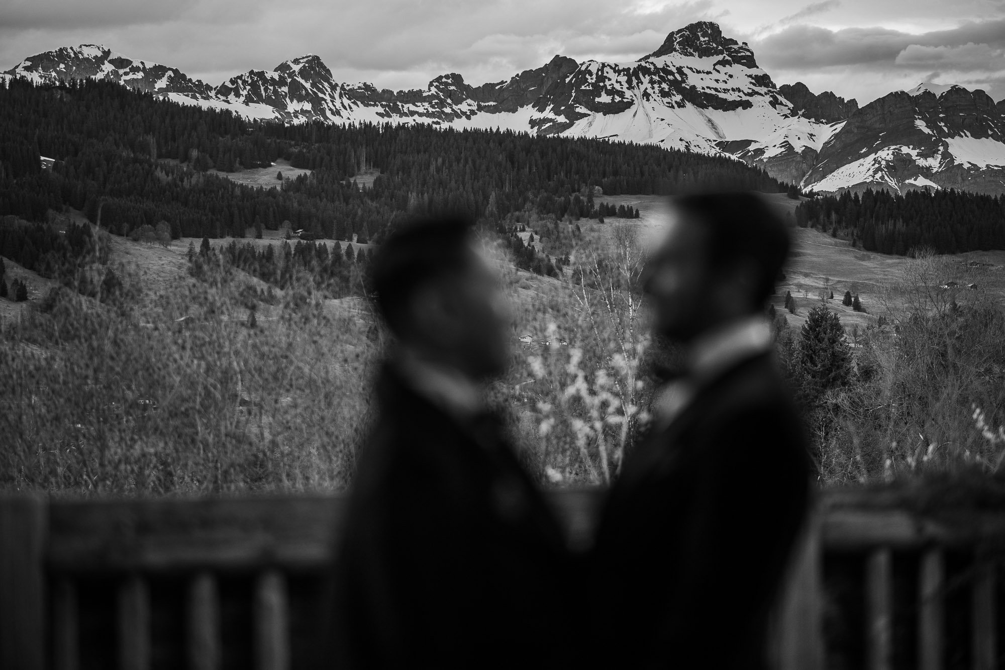 Deux personnes floues face à face sur un balcon avec montagnes enneigées en arrière-plan.