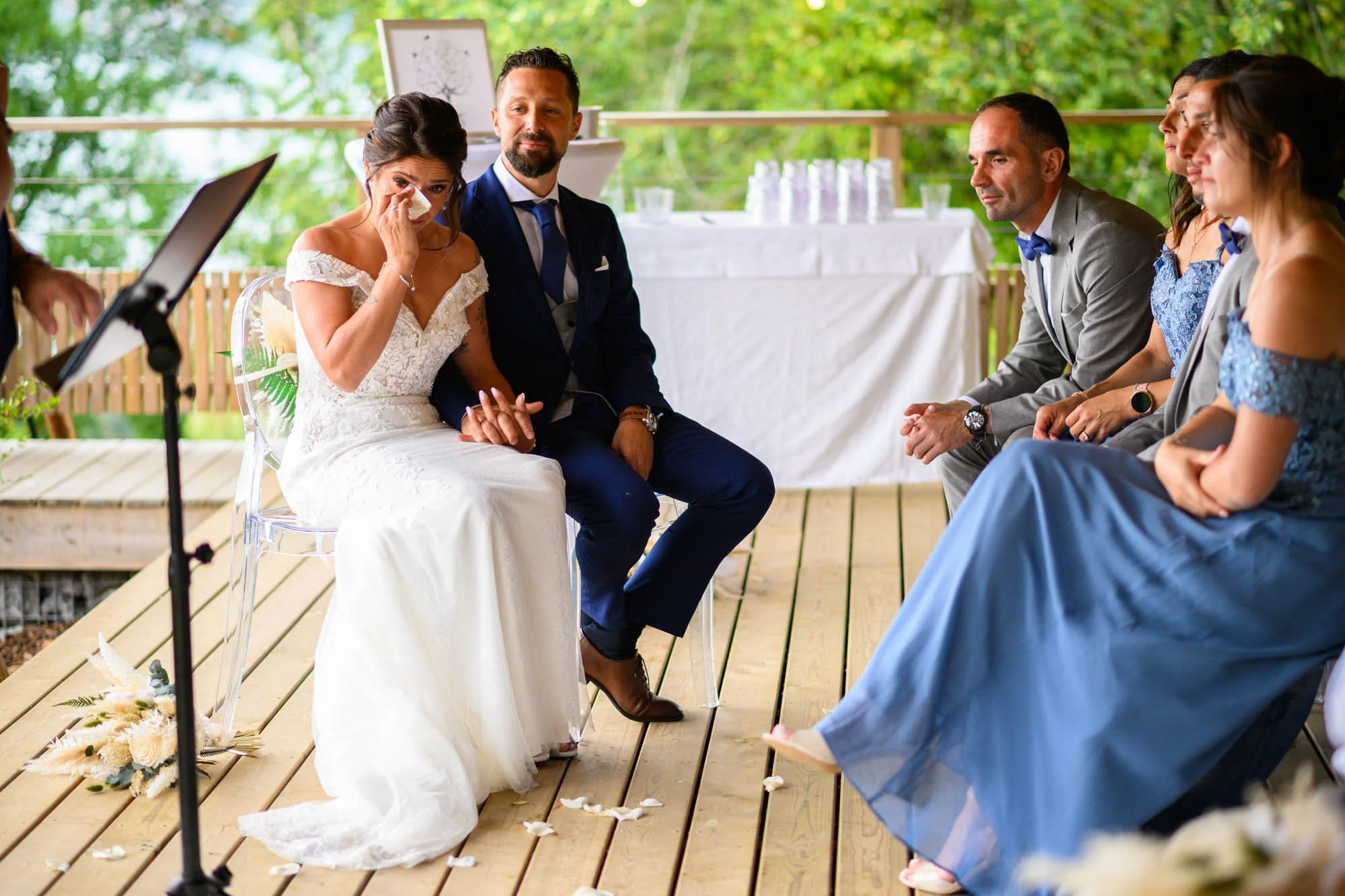 Une femme en robe de mariée pleure en tenant la main d'un homme en costume lors d'une cérémonie de mariage en plein air, avec d'autres invités assis à côté sous un abri en bois dans un environnement verdoyant.