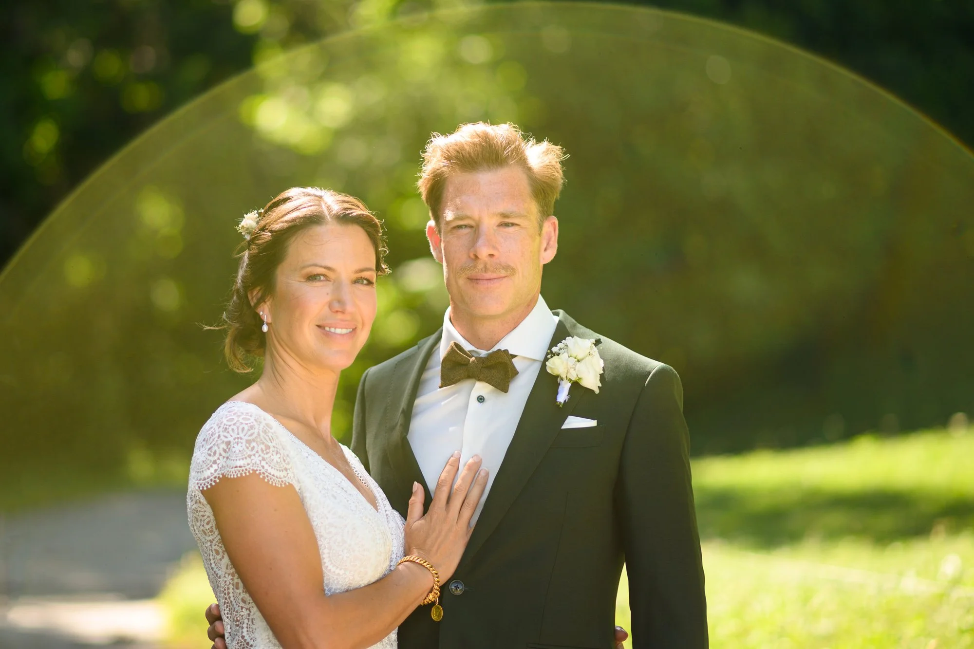 Un couple en costume de mariage pose à l'extérieur avec un fond de nature verdoyante. La femme porte une robe blanche en dentelle et des boucles d'oreilles, tandis que l'homme est en costume noir avec une cravate noire et une boutonnière blanche.