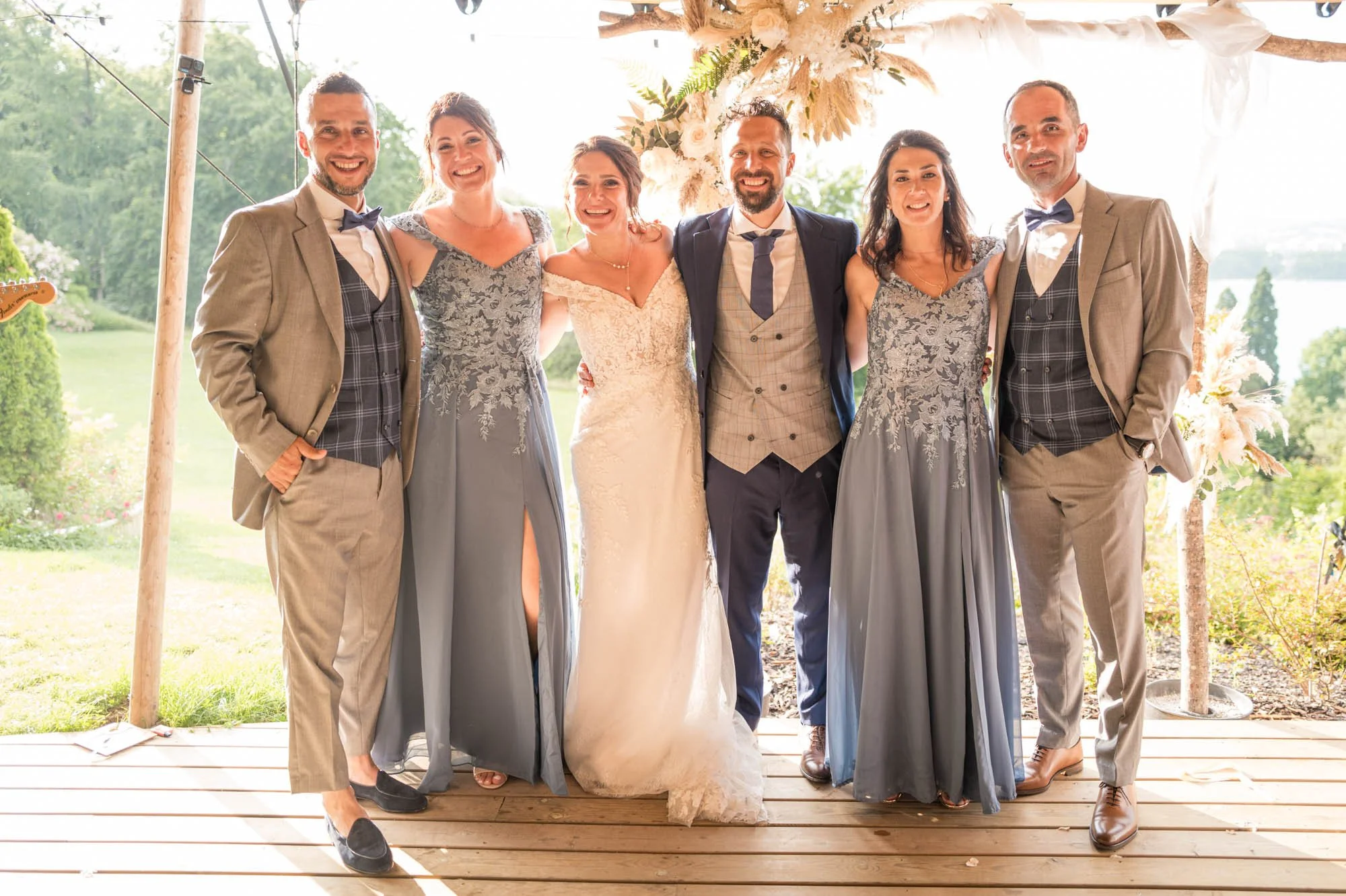 Groupe de personnes lors d'un mariage en plein air, comprenant deux hommes en costumes et quatre femmes en robes élégantes, sous une structure en bois décorée de fleurs, avec un paysage verdoyant en arrière-plan.