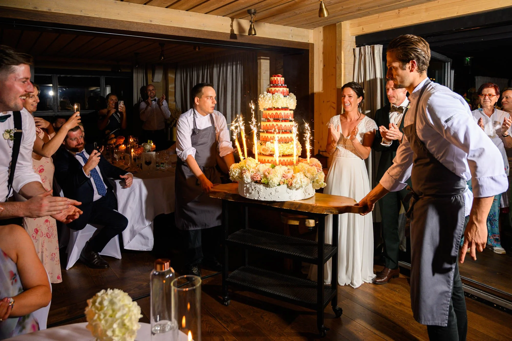 Une scène de mariage où un gâteau de mariage spectaculaire est présenté, entouré de convives qui applaudissent et prennent des photos, dans une salle décorée chaleureusement.