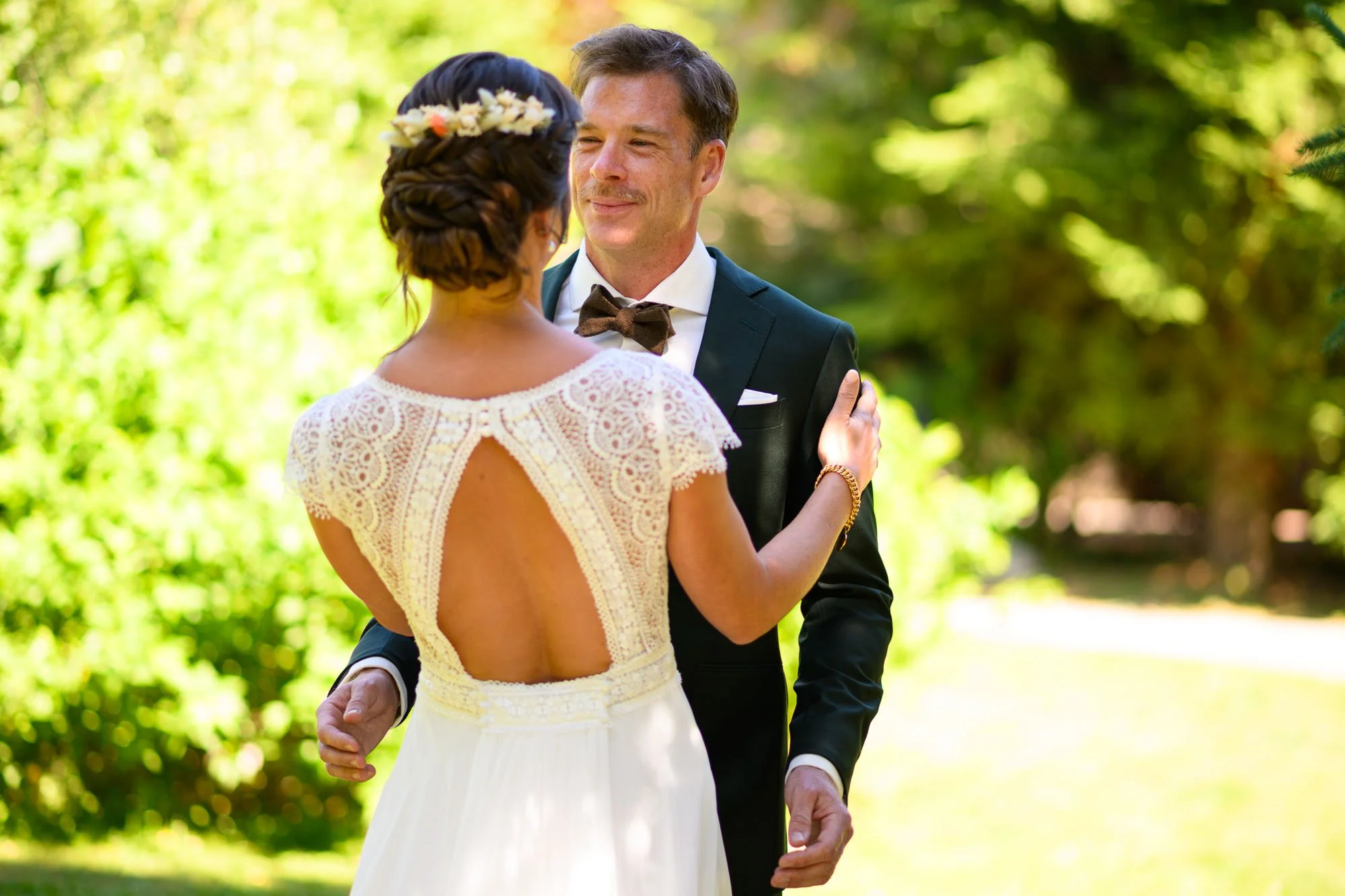 Un couple lors d'un mariage en plein air. La femme porte une robe blanche avec un dos nu, et l'homme porte un costume noir avec un nœud papillon brun, dans un environnement verdoyant