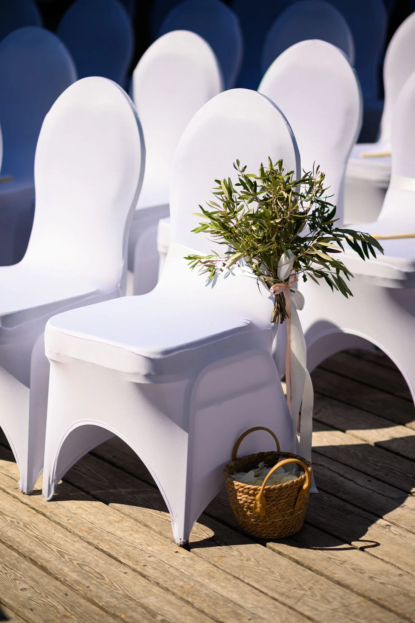 Chaises blanches couvertes de tissu dans une configuration de mariage, avec un bouquet d'oliviers attaché à une chaise et un panier en osier au sol rempli de pétales de fleurs