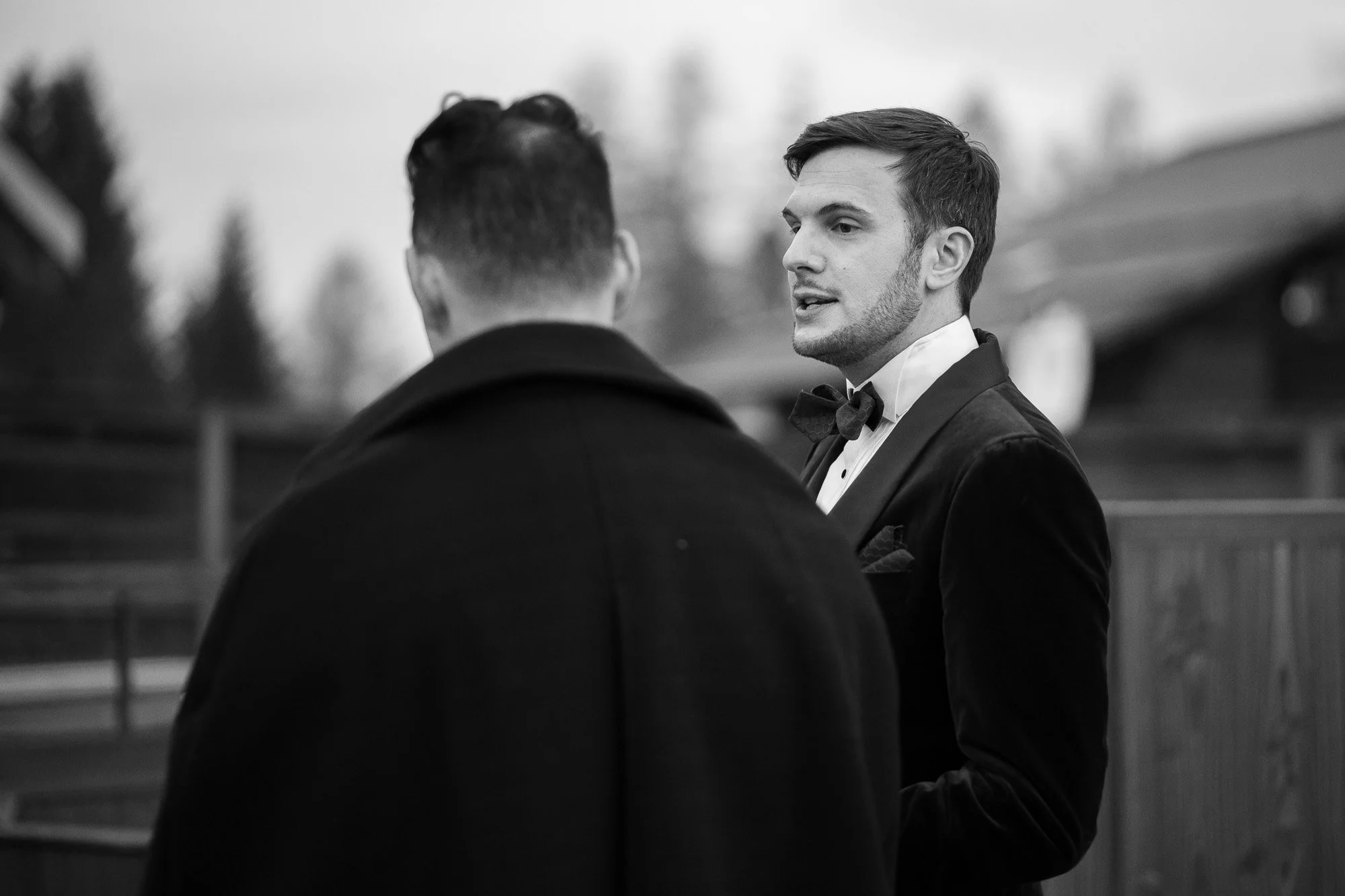 Deux hommes en costume de smoking discutent en extérieur, un en face de l'autre, avec un fond flou de bâtiments et de nature.