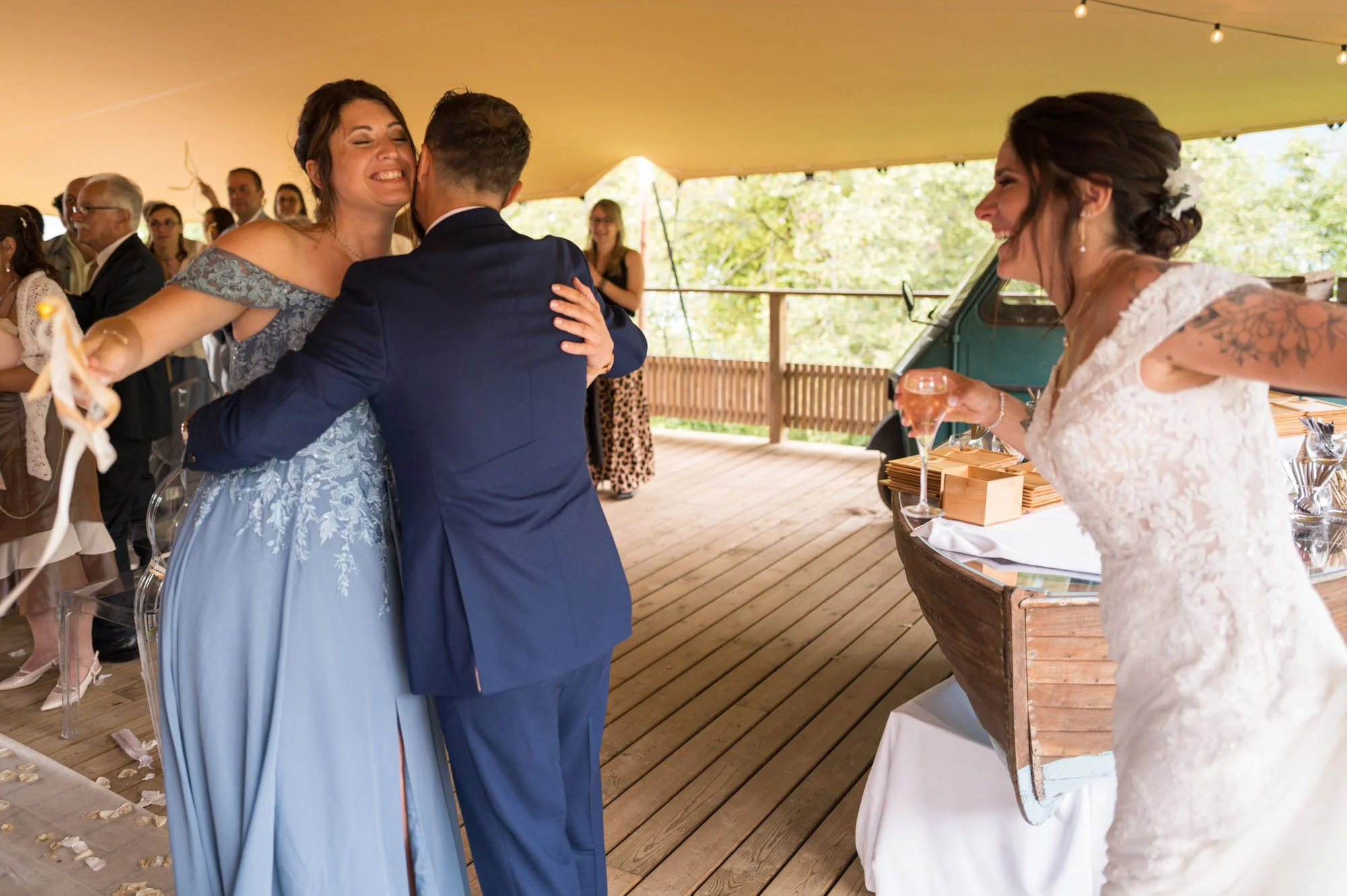 Une mariée en robe blanche et une femme en robe bleue heureuse à un mariage, entourées d'invités.