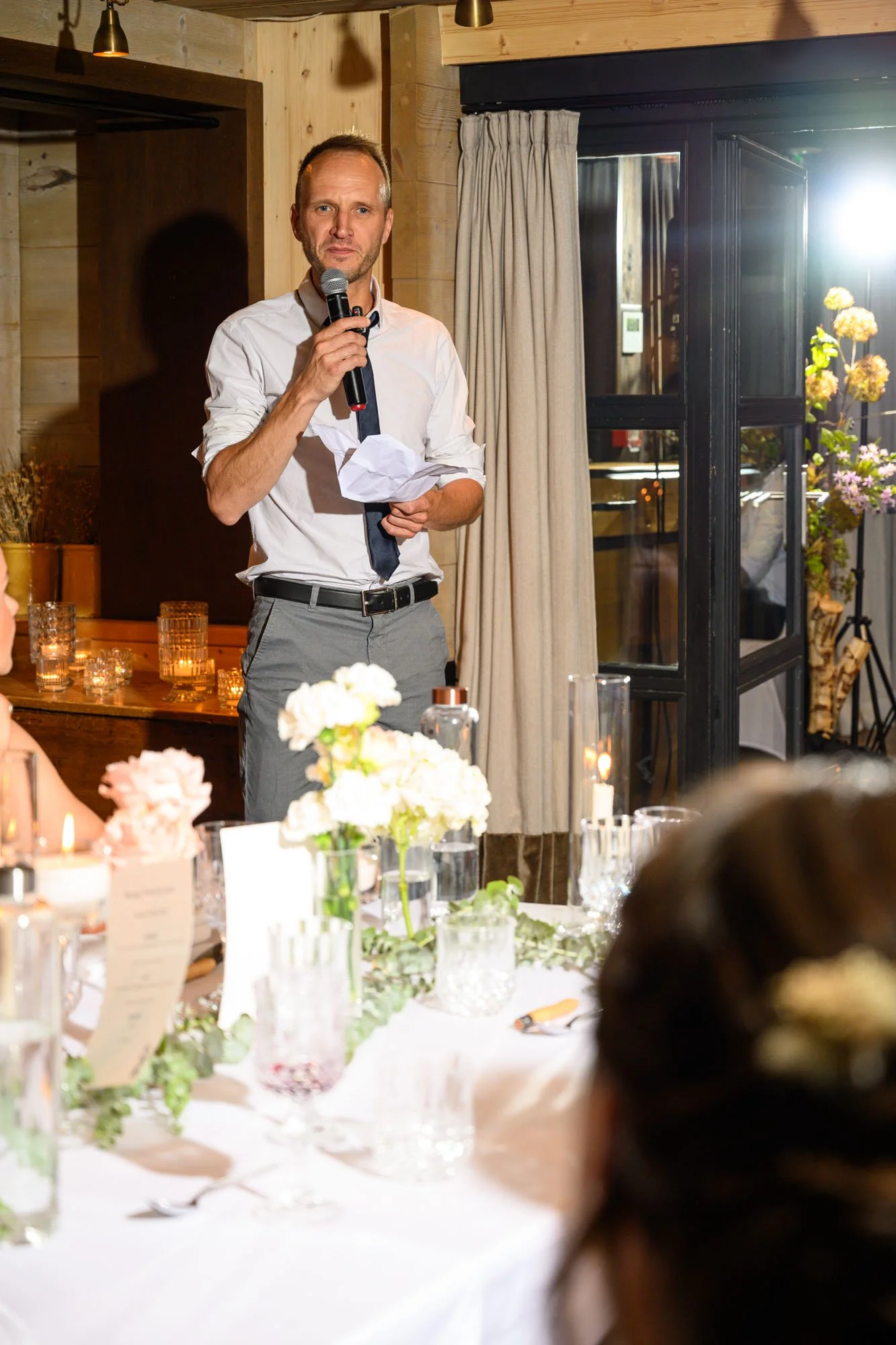 Un homme parle en tenant un micro lors d'un événement dans un cadre chaleureux avec des décorations florales et de bougies.