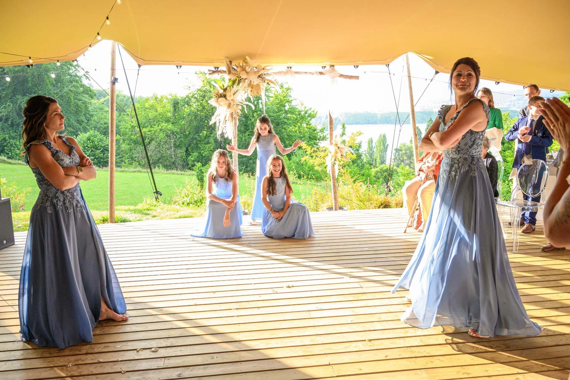 Un groupe de personnes danse lors d'une cérémonie en plein air sous une tente, avec des femmes en robes longues de couleur gris-bleu et des jeunes filles en robes bleues, dans un décor naturel avec verdure et un lac en arrière-plan.
