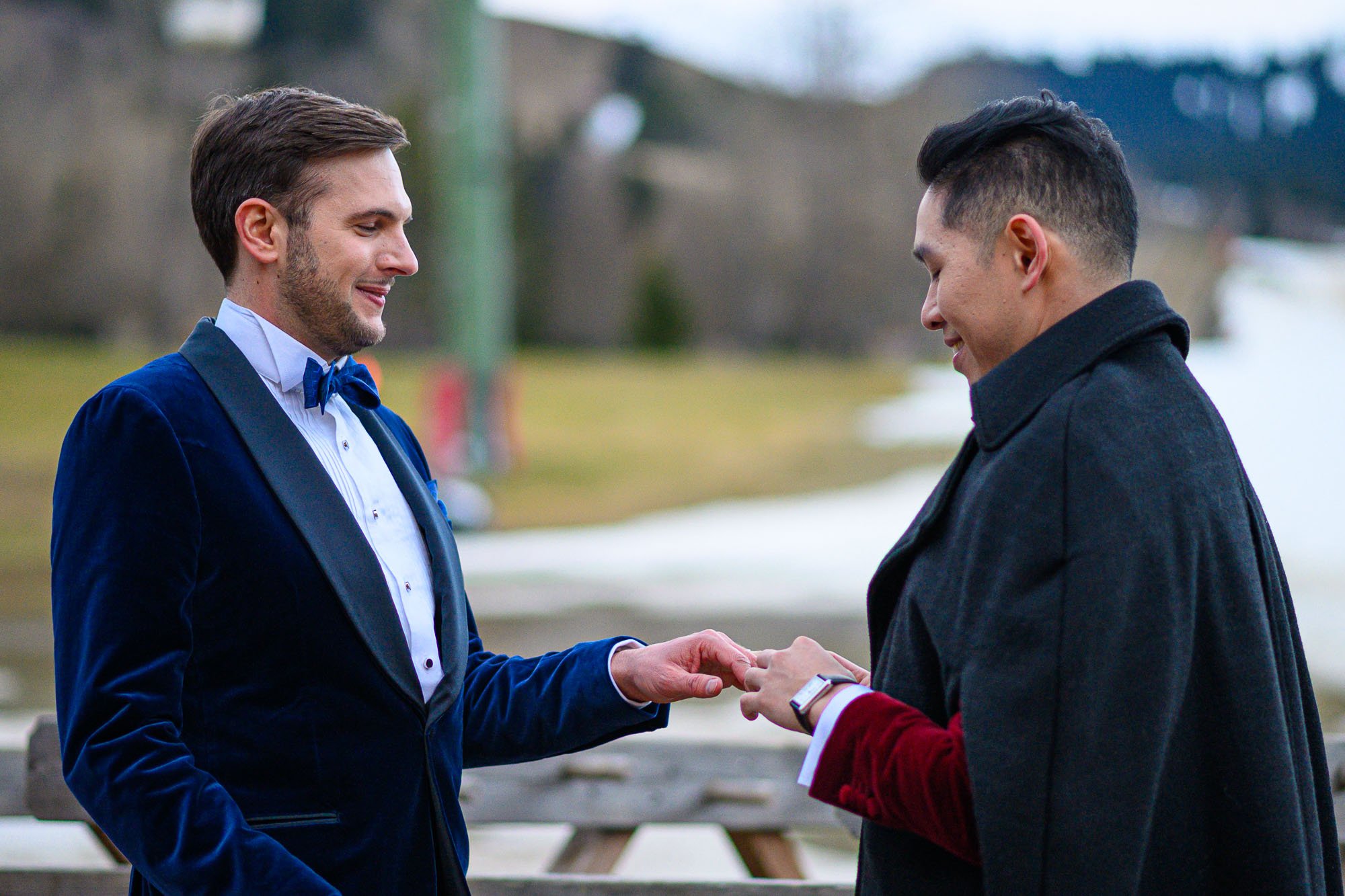 Deux hommes échangent des alliances lors d'une cérémonie de mariage en extérieur, l'un vêtu d'une veste en velours bleu et l'autre d'un manteau noir.