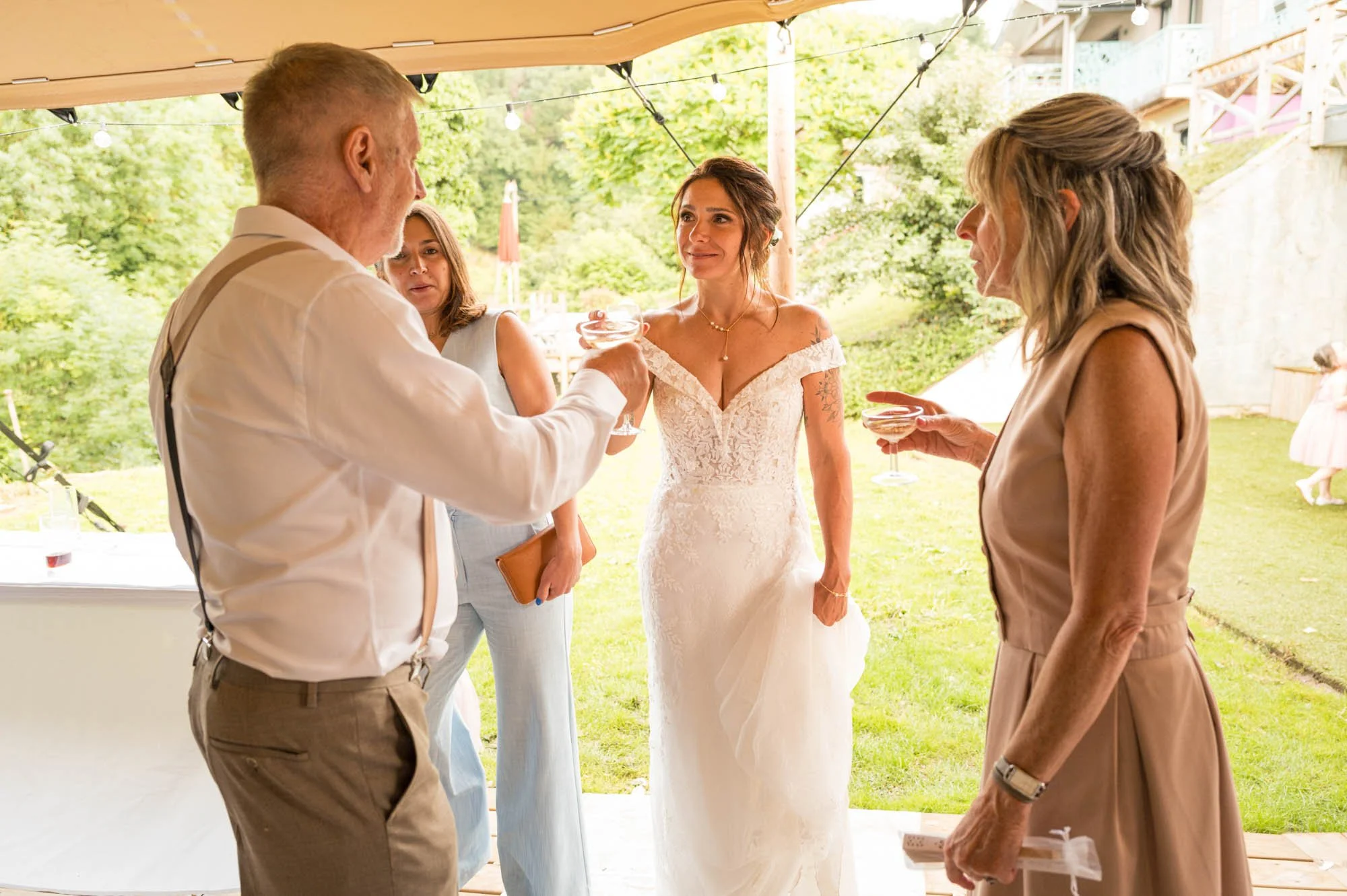 Cinq personnes en costume élégant se tiennent sous une tente, à l'extérieur, en train de trinquer avec des coupes de champagne lors d'une célébration de mariage.