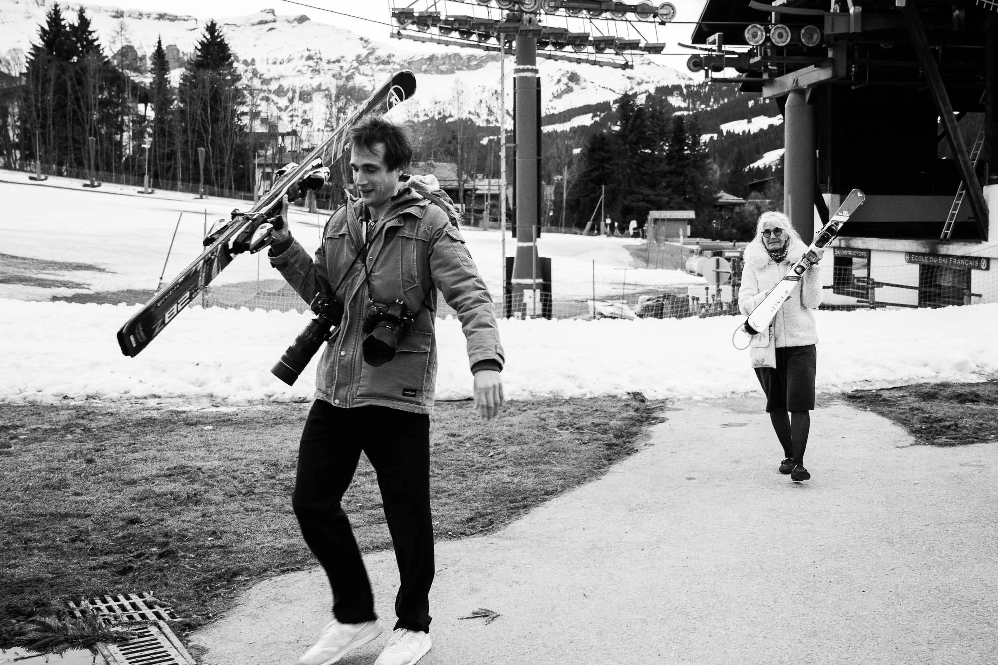 Deux personnes marchent dans une station de ski avec des skis, l'une d'elles tient ses skis sur l'épaule. Le paysage est enneigé avec des montagnes en arrière-plan.