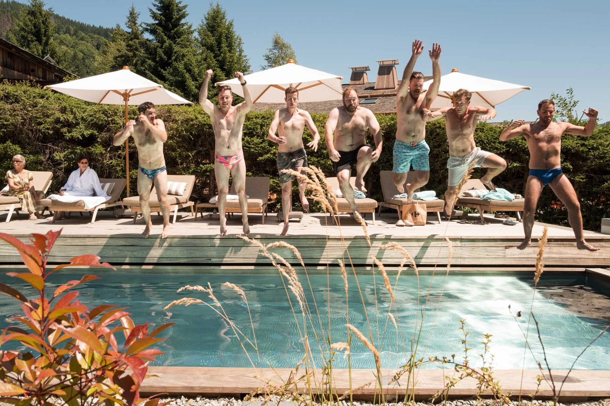 Un groupe d'hommes sautant dans une piscine lors d'une fête au bord d'une piscine, avec des parasols et des personnes assises en arrière-plan.