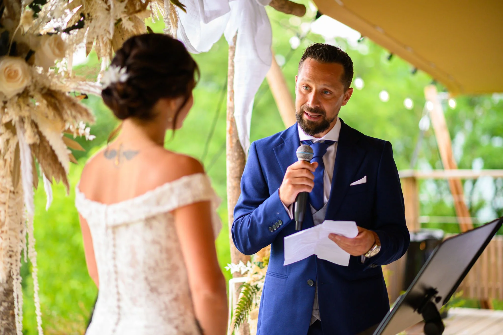 Un homme en costume bleu lit un discours lors d'une cérémonie de mariage, face à une bride en robe blanche dans un cadre extérieur verdoyant.