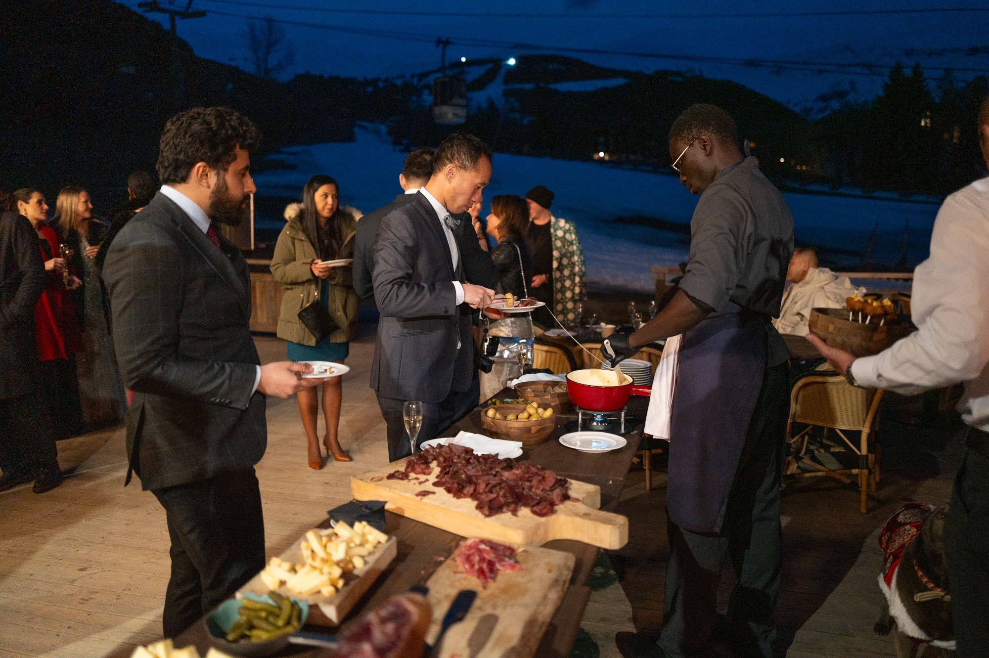 Groupe de personnes en train de servir et de prendre de la nourriture lors d'un repas en extérieur, avec un paysage enneigé en arrière-plan