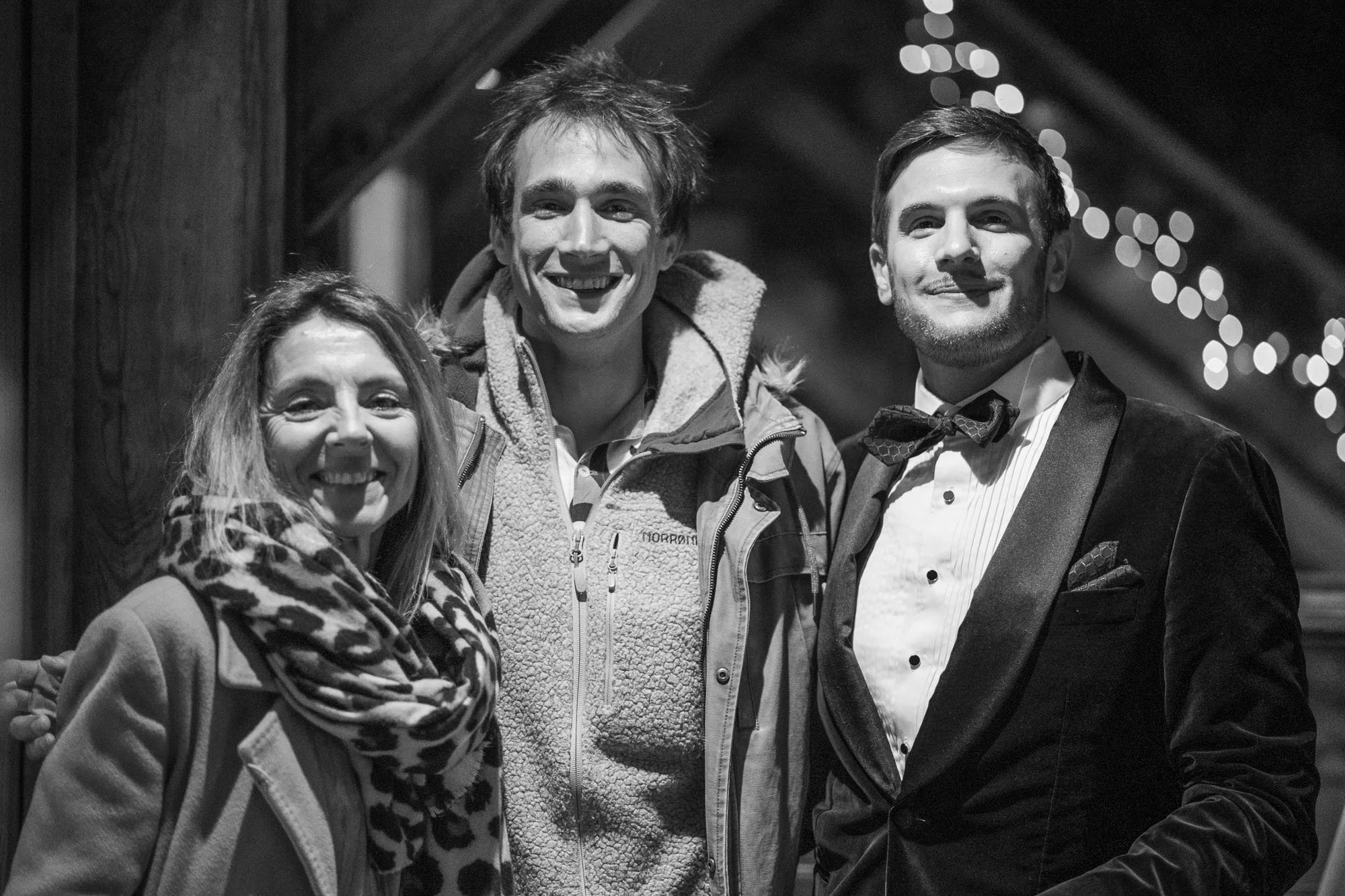 Trois personnes, deux hommes et une femme, posant ensemble en tenue formelle et décontractée, souriant dans un décor festif avec des lumières floues en arrière-plan.