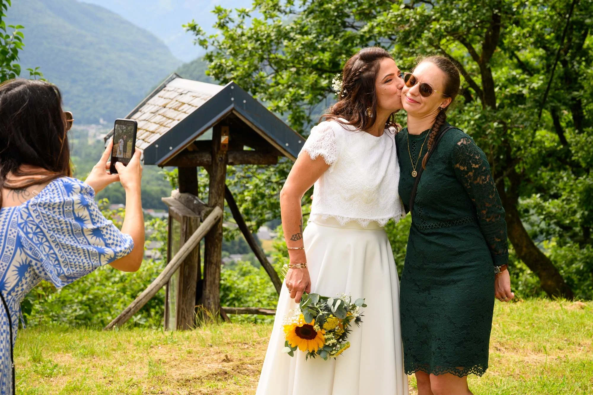 Deux femmes lors d'un mariage, l'une en robe blanche avec bouquet de tournesols, embrasse l'autre en robe noire, pendant qu'une tierce personne prend une photo.